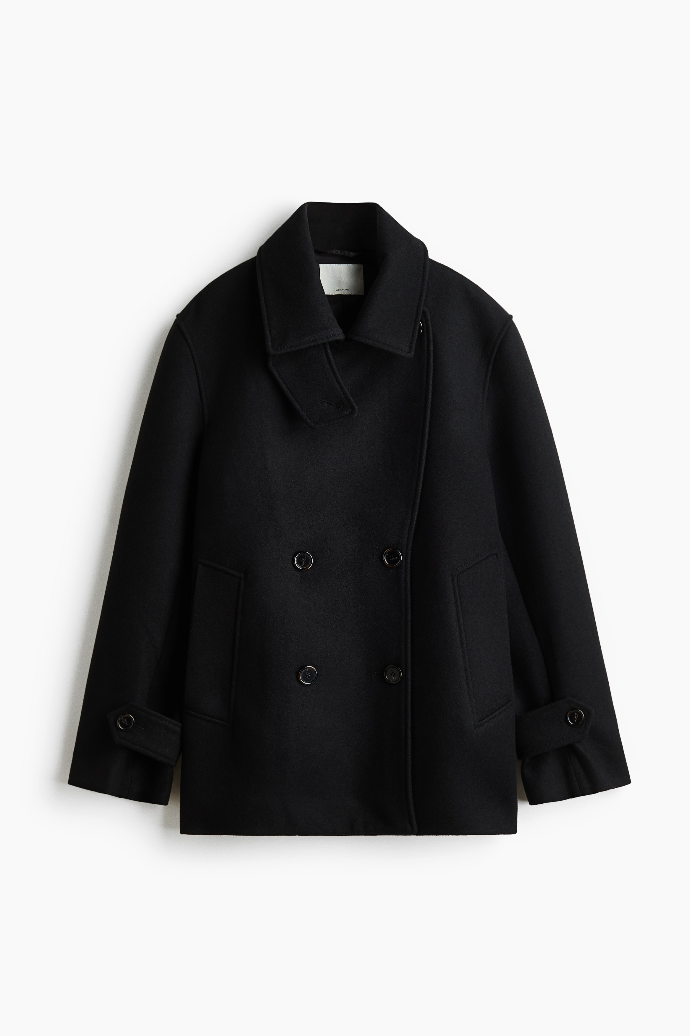 A dark, double-breasted coat featuring dropped shoulders, a wide collar, and buttoned tabs at the cuffs is being worn by the model. The loose-fitting coat is layered open over a white collared shirt, a dark turtleneck, and dark trousers.