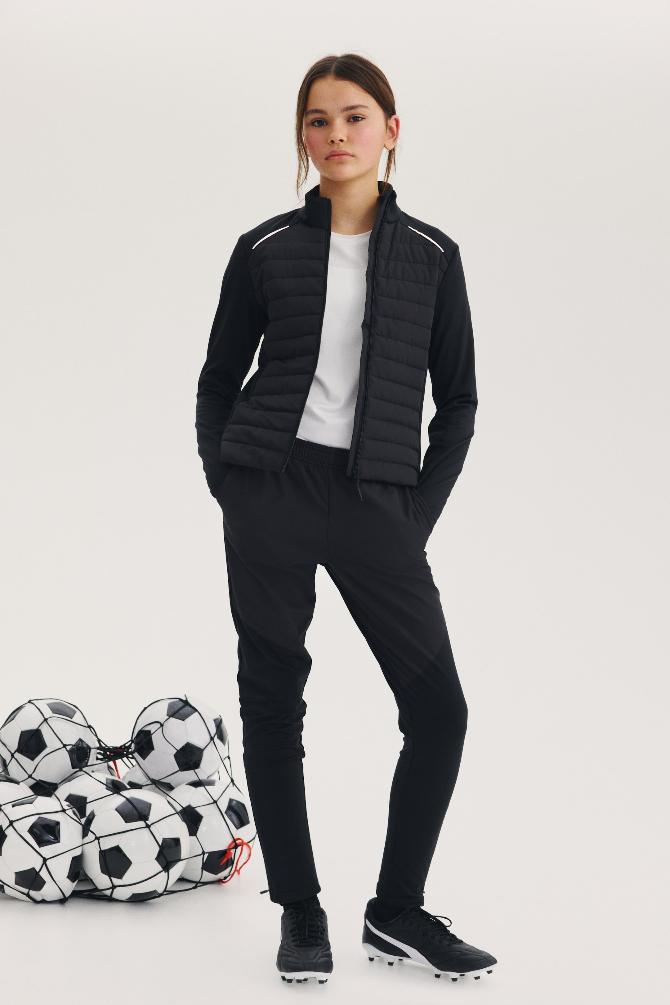 Προβολή μεγαλύτερης εικόνας: Ένα μαύρο αθλητικό μπουφάν με όρθιο γιακά, καπιτονέ εμπρόσθιο πάνελ, λεία μαύρα μανίκια και λευκές ανακλαστικές ρίγες στους ώμους φοριέται πάνω από ένα λευκό T-shirt. Συνδυάζεται με μαύρο αθλητικό παντελόνι με κωνική γραμμή και λεπτομέρειες πάνελ στα κάτω μέρη των ποδιών, καθώς και με μαύρα ποδοσφαιρικά παπούτσια με λευκές πλαϊνές ρίγες.