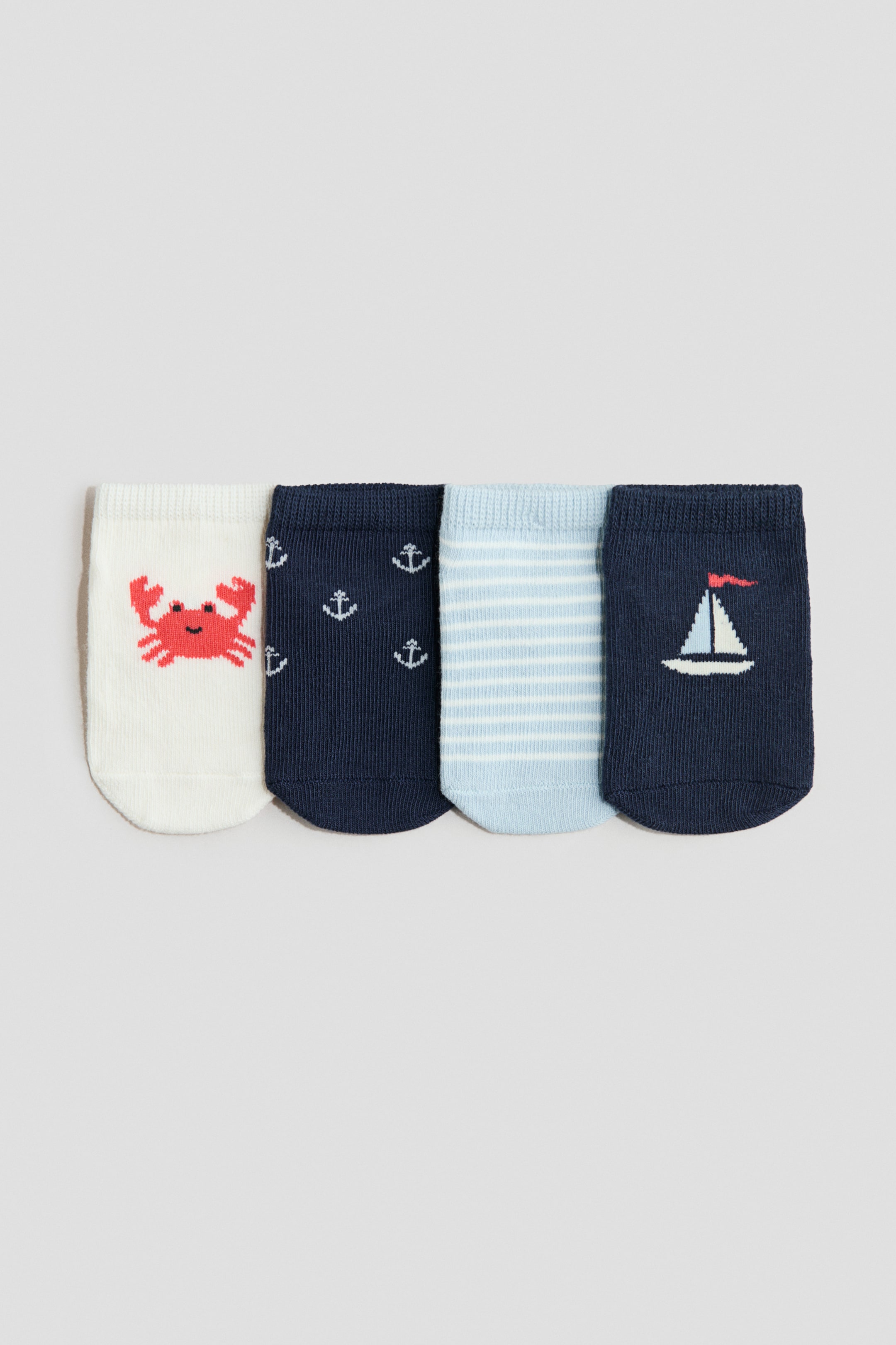 Agrandir l'image: Quatre chaussettes sans couture, en maille fine, avec bords élastiqués sont présentées de face. Une chaussette crème arbore un motif de crabe rouge, une chaussette bleu marine arbore des motifs d'ancre blancs, une chaussette bleu clair arbore des rayures horizontales blanches, et une chaussette bleu marine foncé arbore un motif de voilier blanc avec un drapeau rouge.