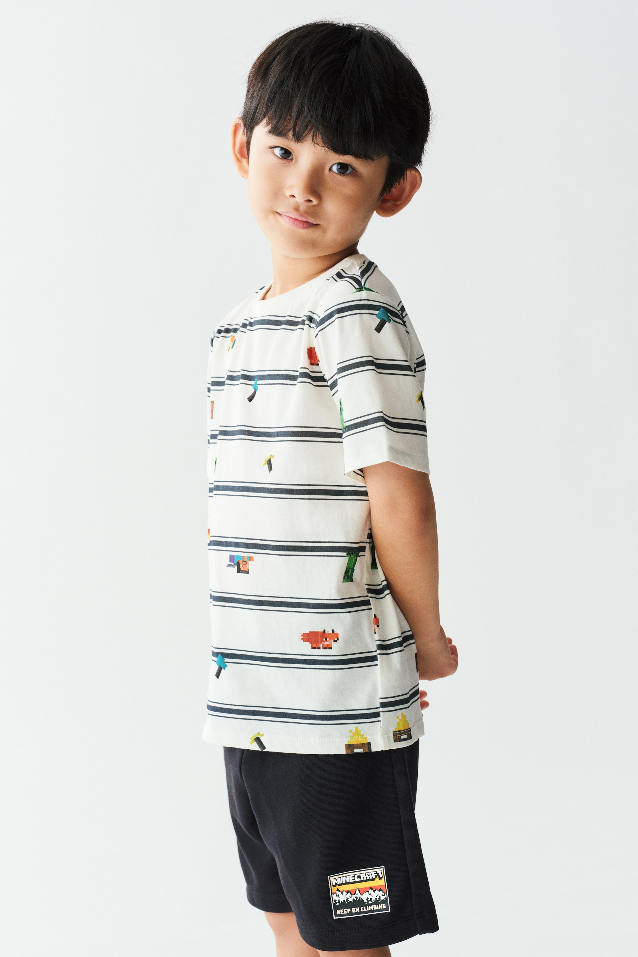 View larger image: A short-sleeved white T-shirt featuring horizontal black stripes and small, colourful pixelated graphics is worn by the model. The tee features a round, rib-trimmed neckline and is paired with matching black shorts.