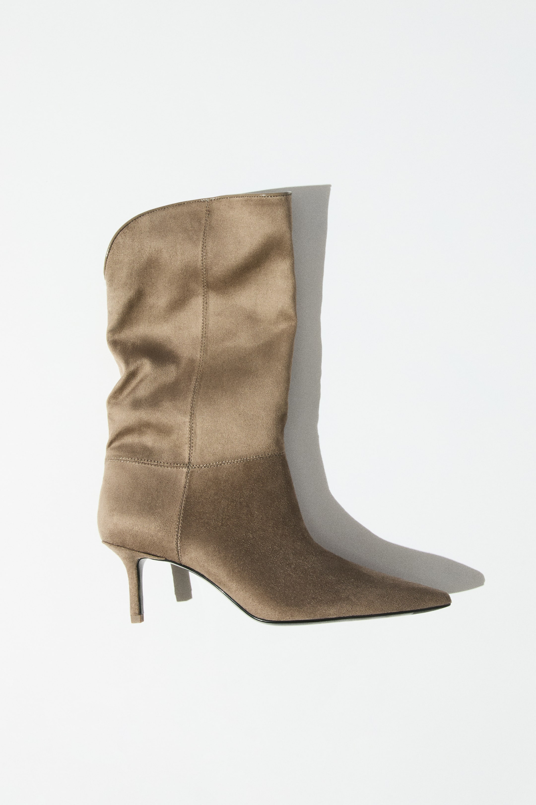 Agrandir l'image: Une paire de bottes taupe à finition douce et veloutée est présentée de profil, tournée vers la droite. Le modèle présente des bouts pointus, des talons étroits et des tiges souples montant jusqu’au mollet avec une ligne supérieure incurvée.