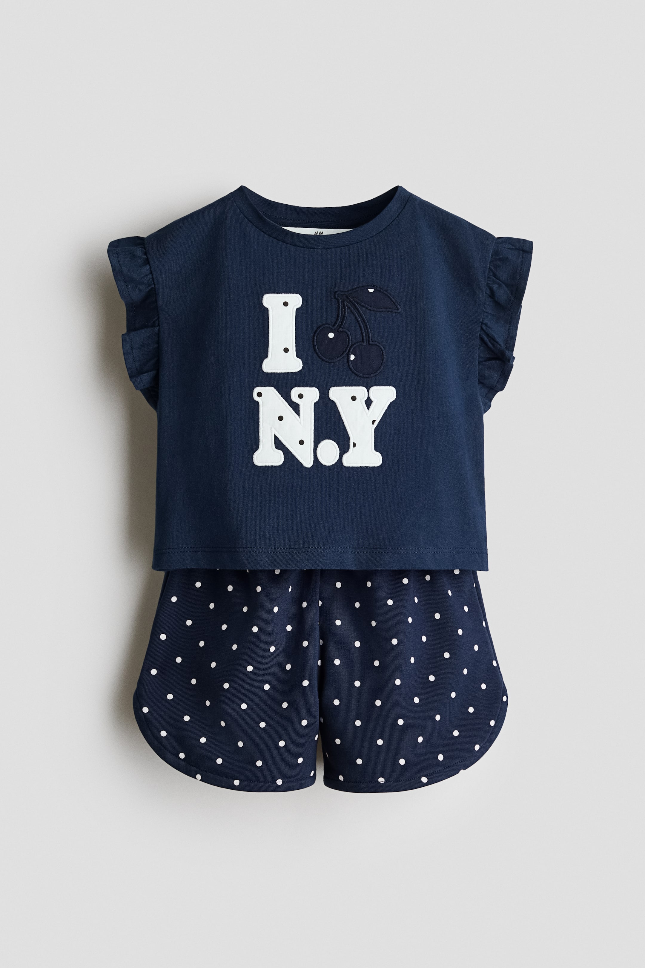 Agrandir l'image: Un ensemble de pyjama deux pièces bleu foncé comprenant un haut et un short. Le haut arbore l'inscription "I [icône de cerise] N.Y." en appliqué blanc avec une cerise brodée bleu foncé, et des manches à volants. Le short assorti présente un motif à pois blancs et des ourlets arrondis.