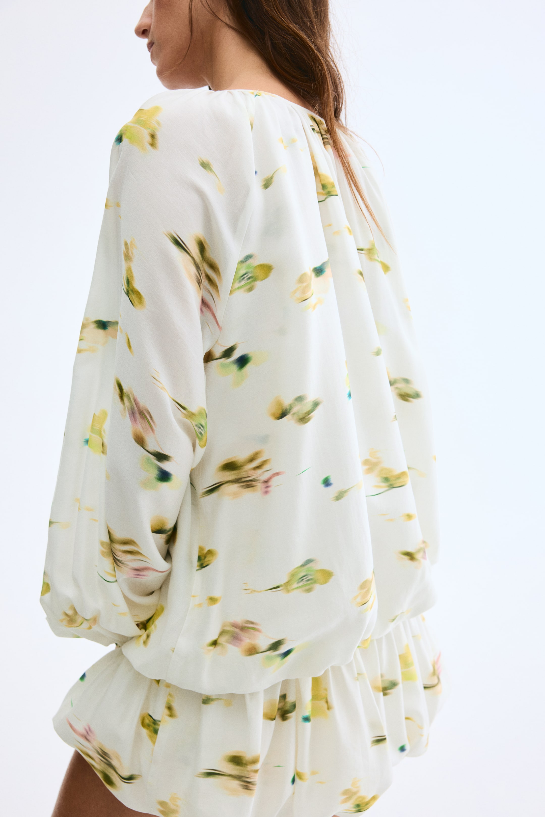 Ampliar la imagen: Un vestido corto de tejido de viscosa blanca, con un patrón abstracto en amarillo y verde, está siendo llevado por la modelo. Luce un escote redondo fruncido, mangas largas raglán tipo globo y una falda de dobladillo abombado, destacando su silueta volumétrica.