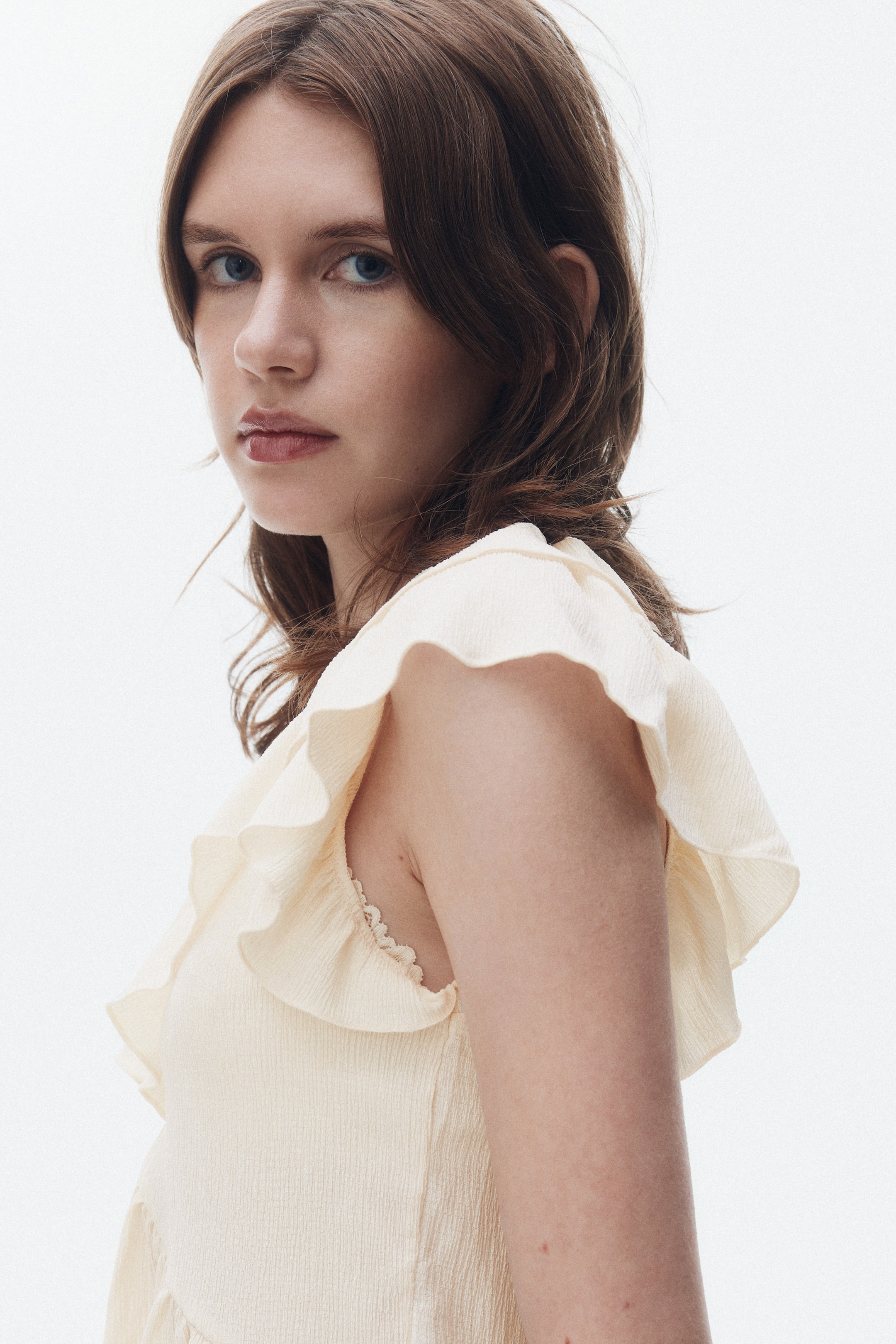 Ampliar la imagen: Un top de color crema claro, visto desde un ángulo frontal lateral, presenta una tela texturizada. Los volantes anchos detallan los hombros y un delicado encaje se puede ver debajo de los volantes.