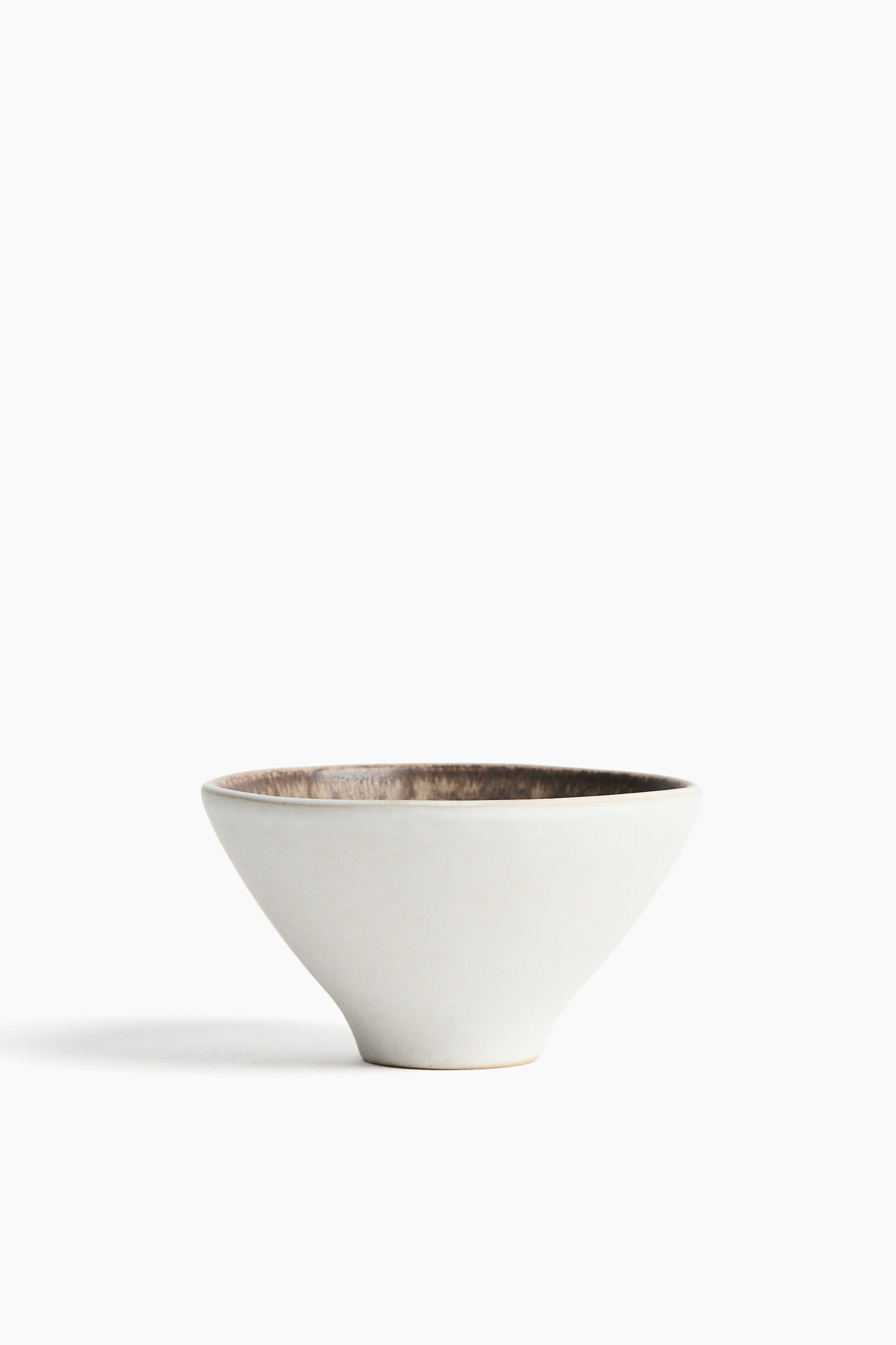 Zobacz większy obraz: Ceramiczna miska stoi pionowo, prezentując zwężającą się ku dołowi stożkową sylwetkę, która przechodzi w niewielką, podstawę na stopce. Zewnętrzna powierzchnia pokryta jest matowym, złamanym białym szkliwem, natomiast wewnętrzny brzeg odsłania kontrastujące ciemnobrązowe, teksturowane szkliwo.