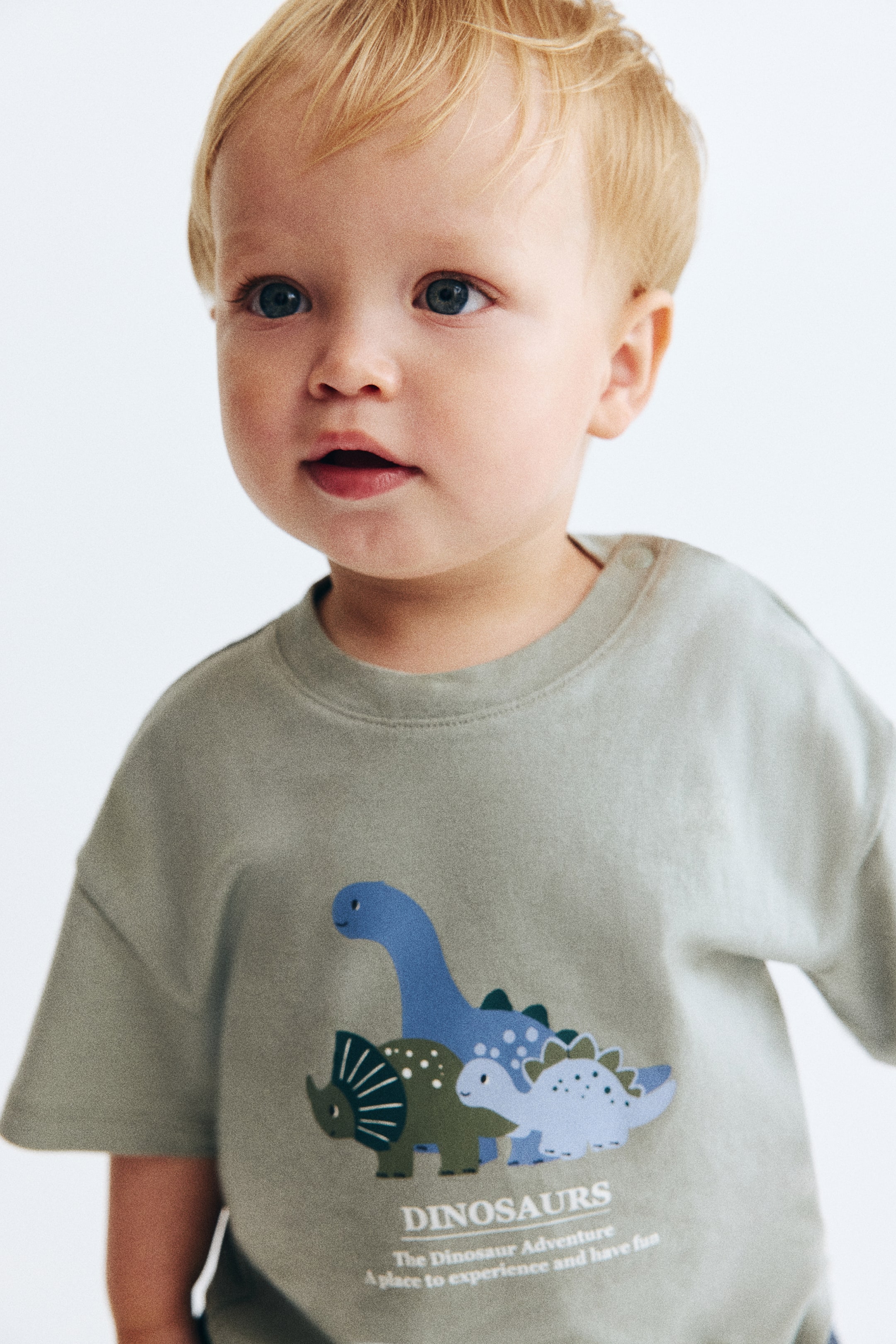 View larger image: A light olive green cotton T-shirt featuring a crew neck with snap buttons on the shoulder and short sleeves. The front showcases a vibrant graphic print of cartoon dinosaurs along with accompanying text.