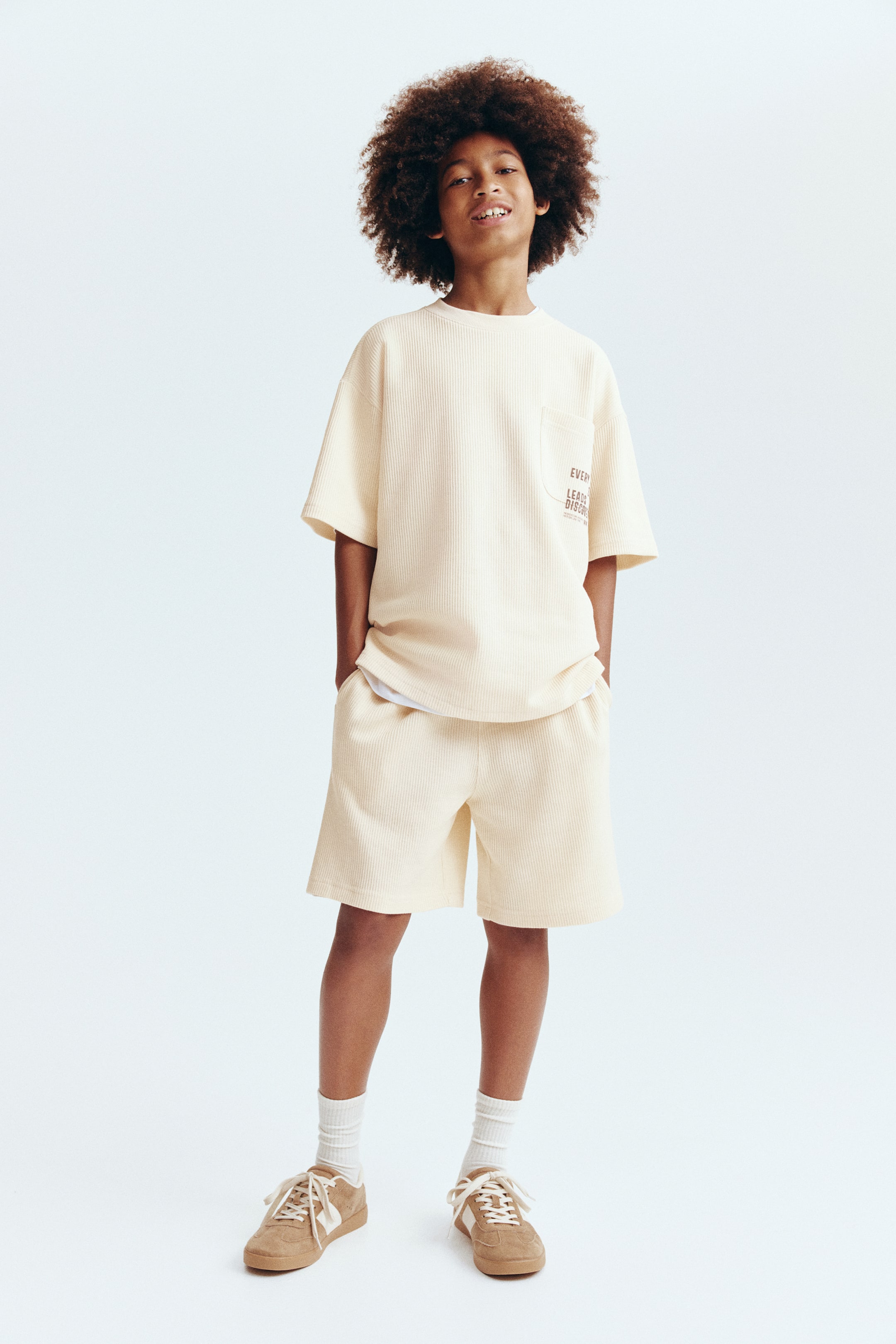 View larger image: A light yellow ribbed knit t-shirt and matching shorts set is being worn, facing forward. The oversized t-shirt features a round neck, short sleeves, and a left chest patch pocket displaying partial brown text, with a white undershirt peeking from below its hem. The mid-thigh length shorts include side pockets.