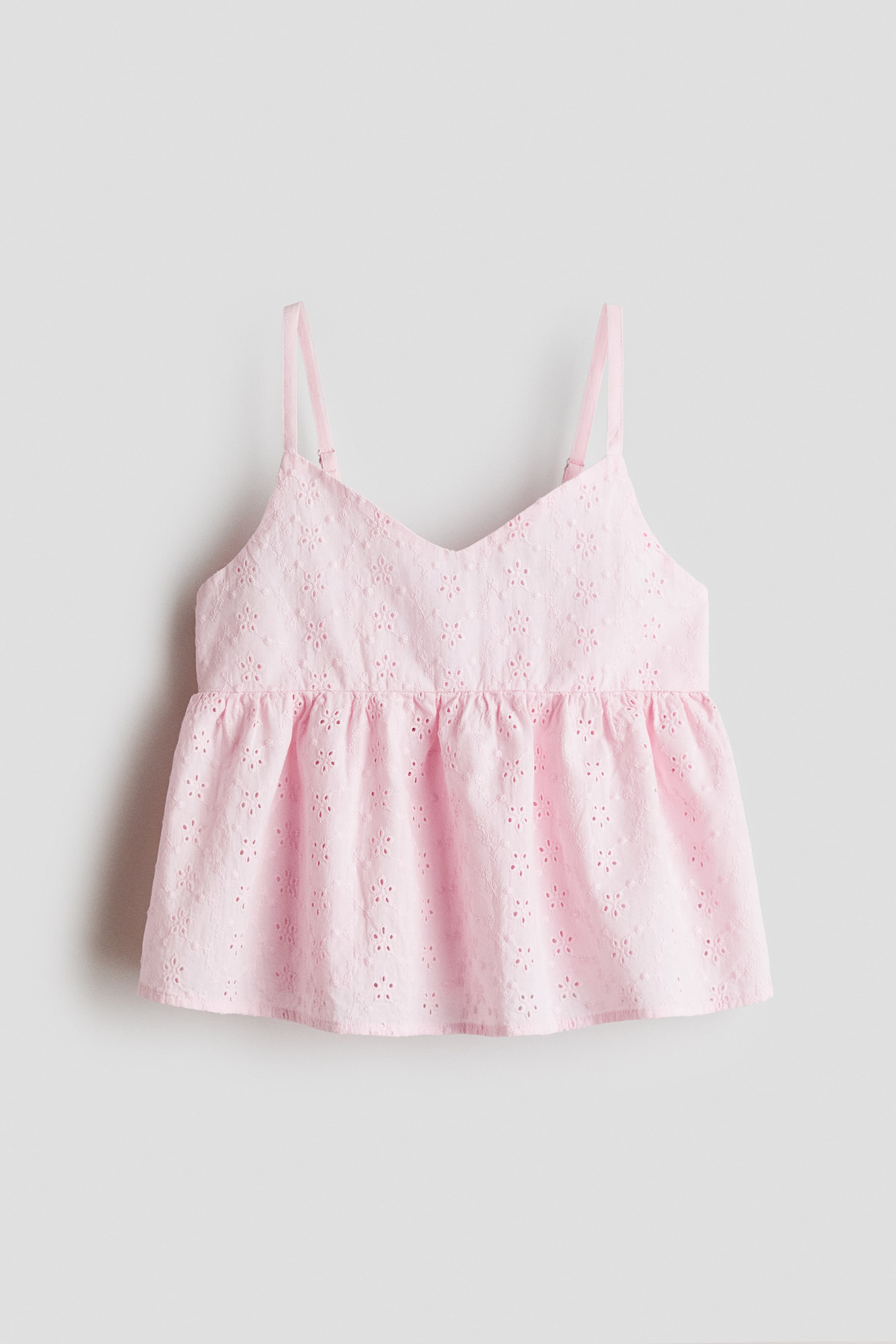 View larger image: A pink top is showcasing its front view, crafted from a cotton weave with embroidered cutouts, featuring a V-shaped neckline and narrow, adjustable shoulder straps. A gathered seam is visible at the waist, leading to a gently flared peplum.
