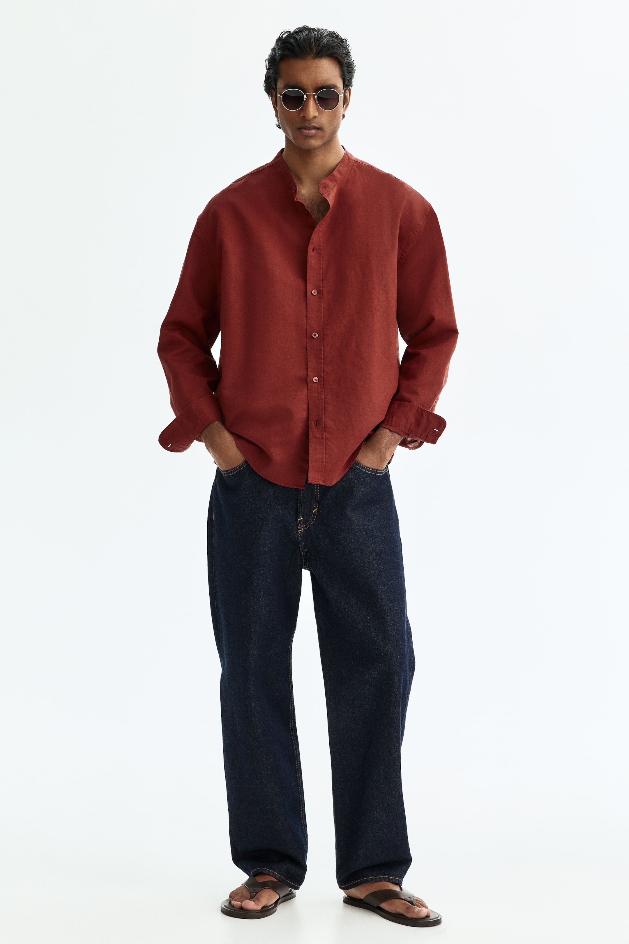 View larger image: A rust-coloured linen shirt is showcasing a band collar and a button-up front. It is featuring long sleeves with rolled-up cuffs and a relaxed, flowing fit.