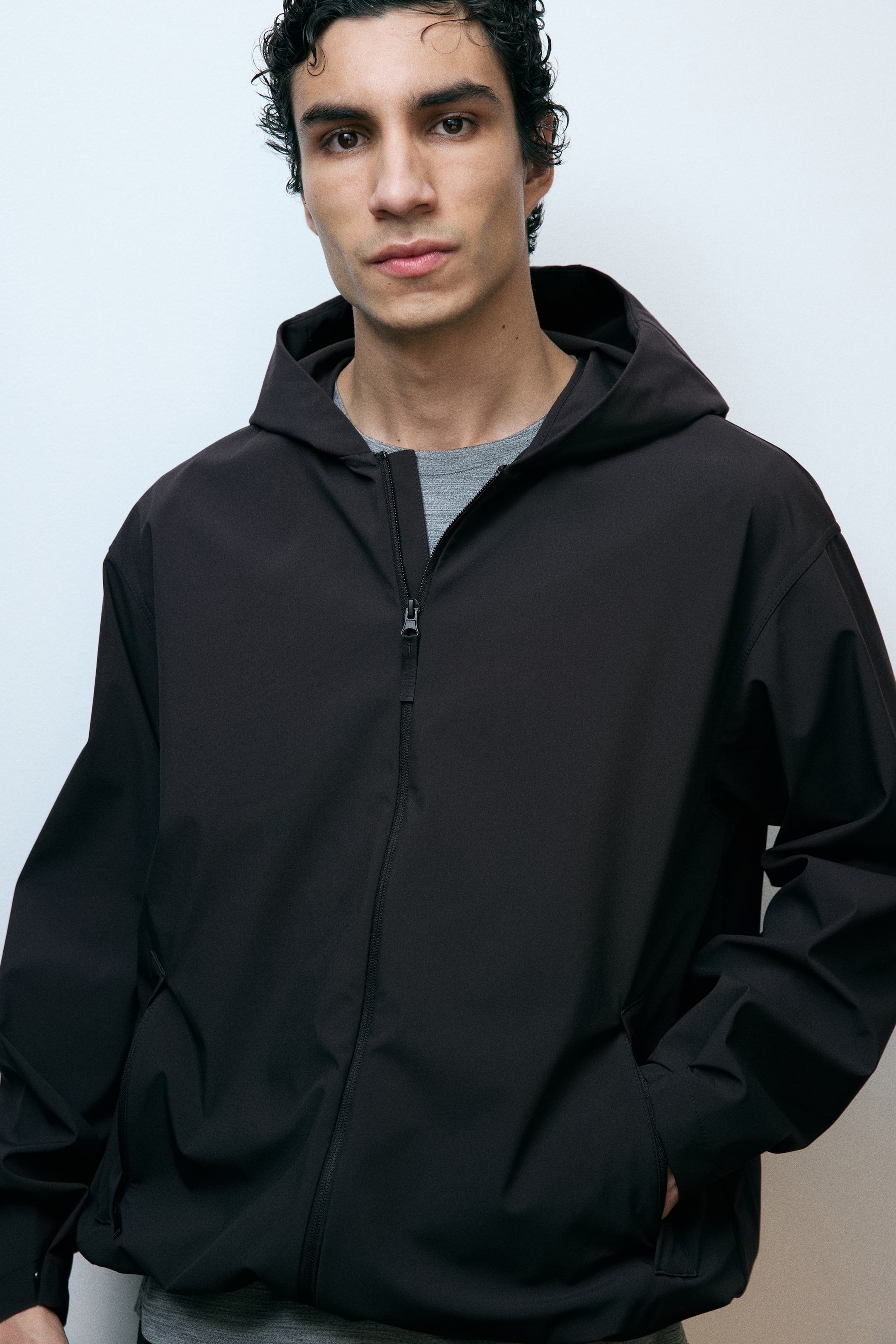 View larger image: The model is wearing a dark sports jacket featuring StormMove?. This hooded jacket is partially zipped, with the model's hand resting in one of its side pockets over a visible gray t-shirt.