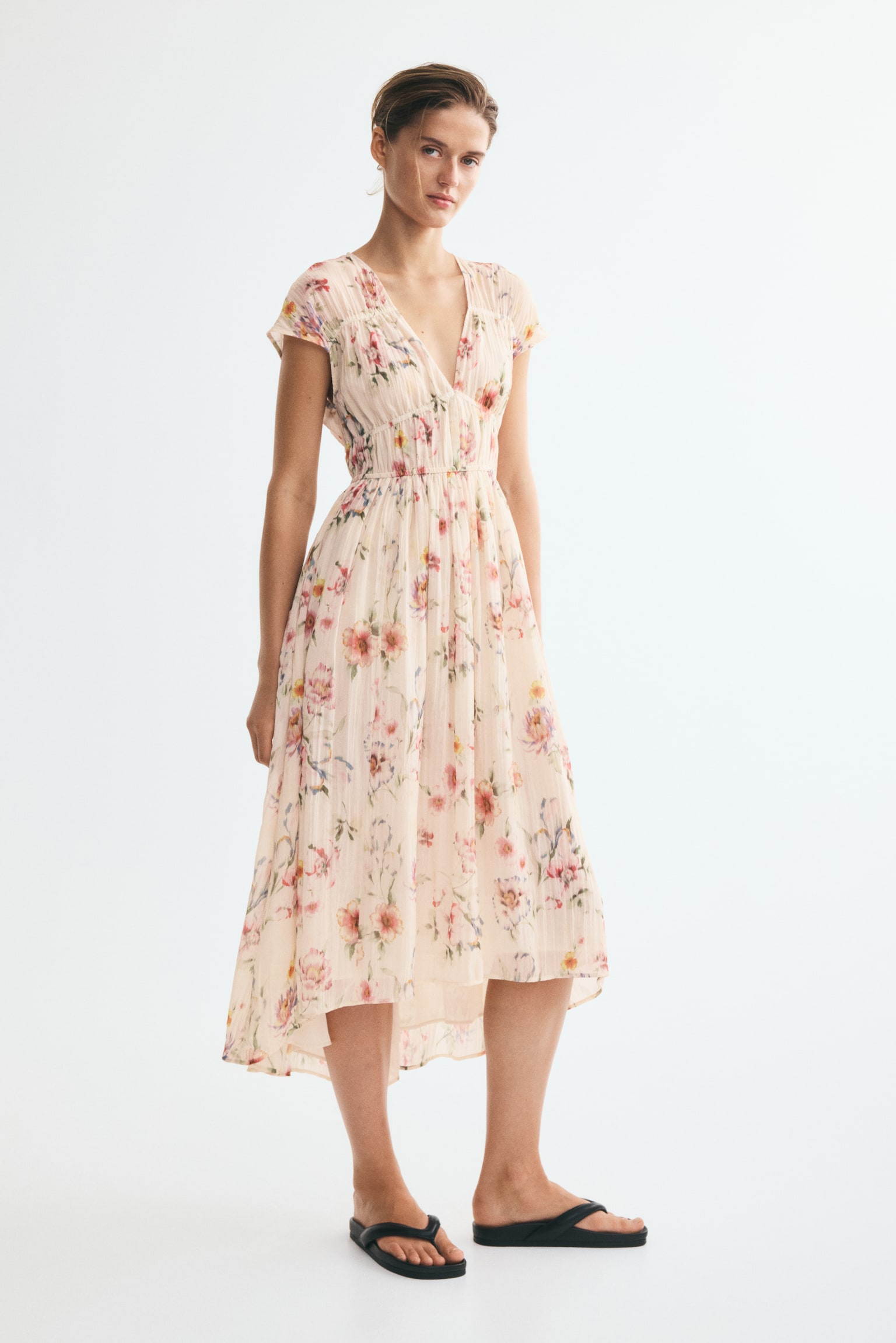 La robe à imprimé floral beige clair présente un décolleté en V, des manches courtes à capuchon et une taille élastiquée froncée, avec sa jupe texturée affichant un ourlet asymétrique et une doublure visible. - 1