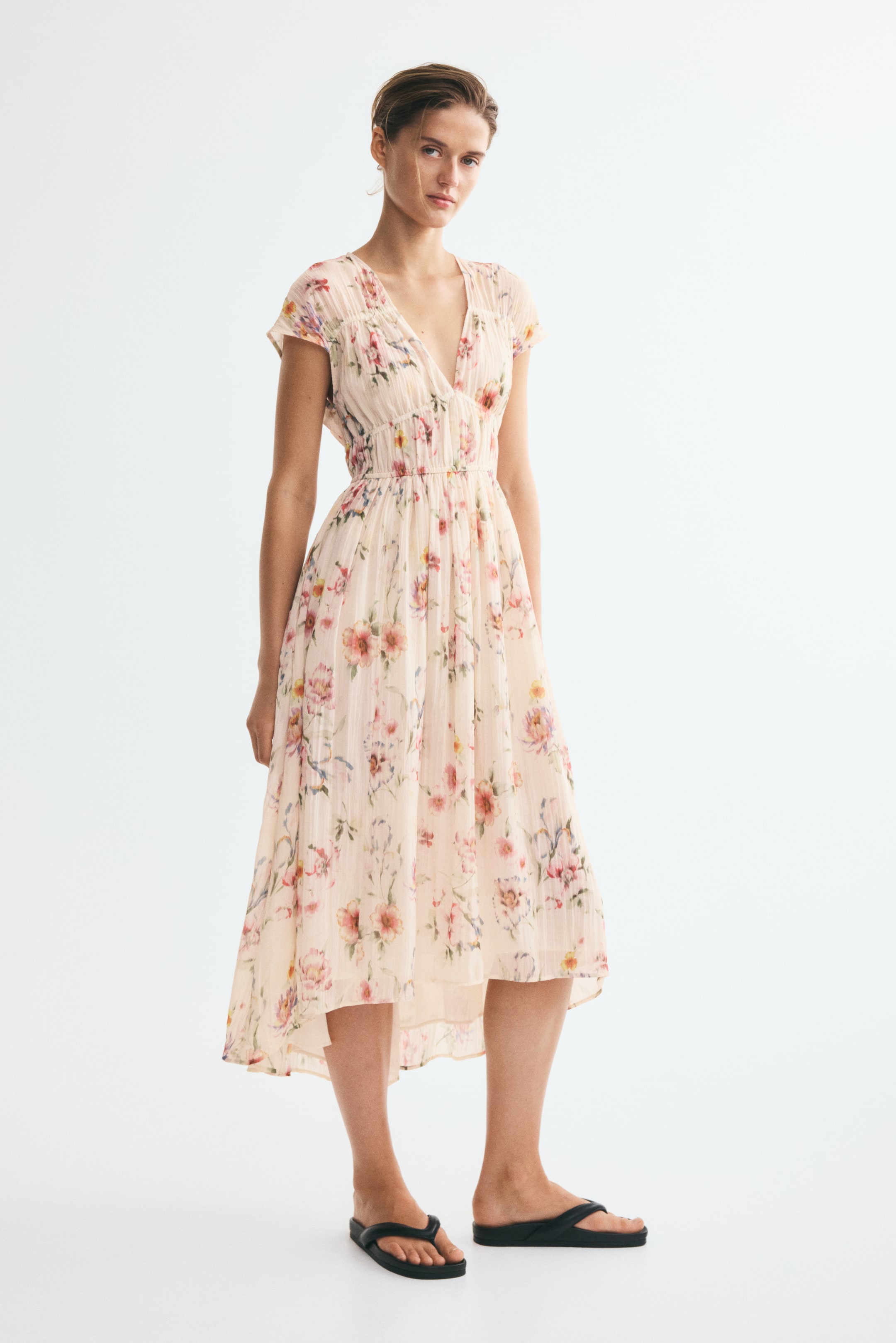 Visa större bild: En vristlång blommig klänning i ett ljust rosa vävt tyg bärs av modellen. Klänningen har V-ringning, korta ärmar, ett rynkat liv och en lätt utsvängd high-low-kjol, kombinerad med svarta slidesandaler.