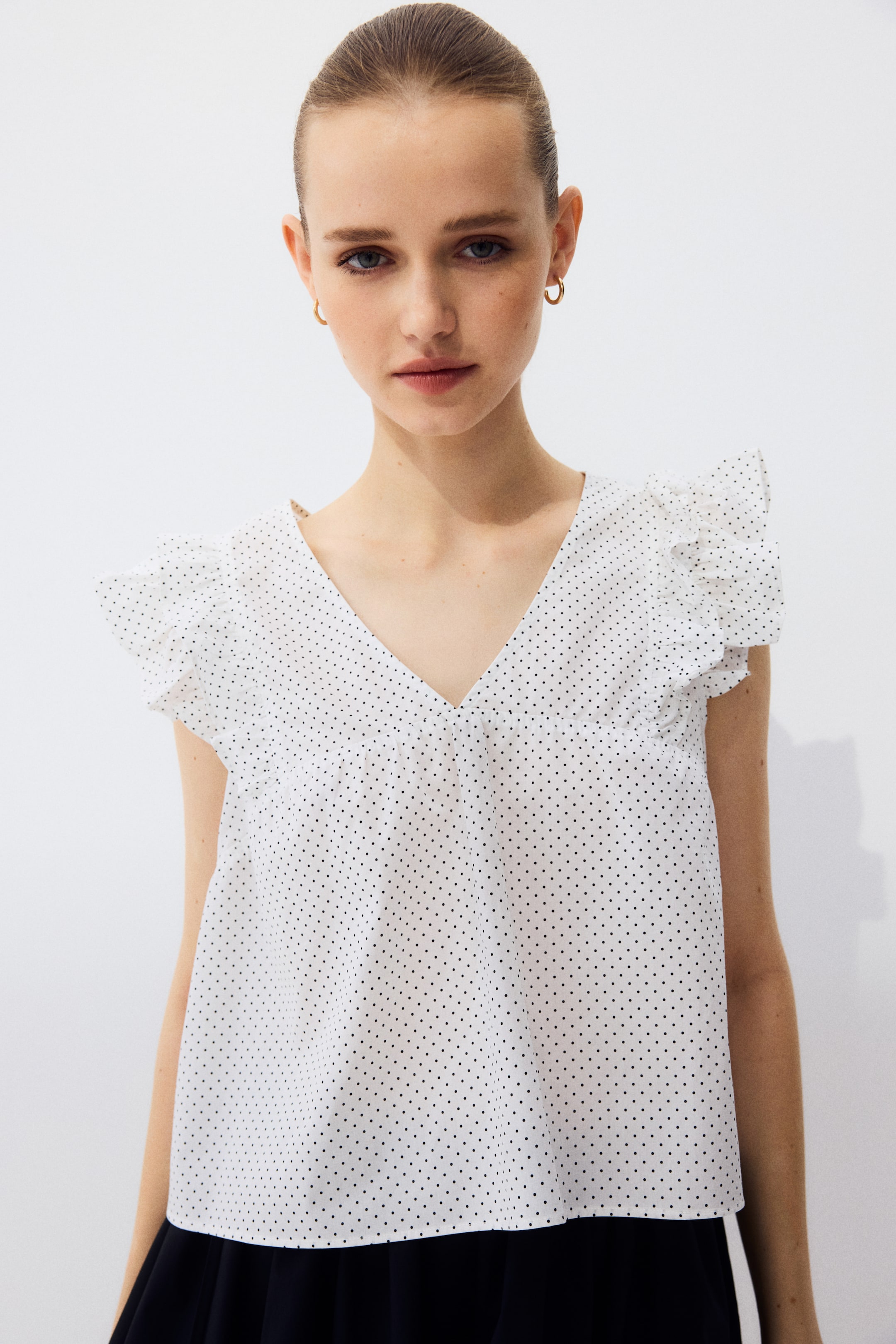 查看大圖: 模特穿著一件白色清爽的棉質波普林上衣，上面有黑色波點，特色包括V字領口、短飄袖、從前面延伸至肩膀的褶邊裝飾，以及聚攏的前幅。