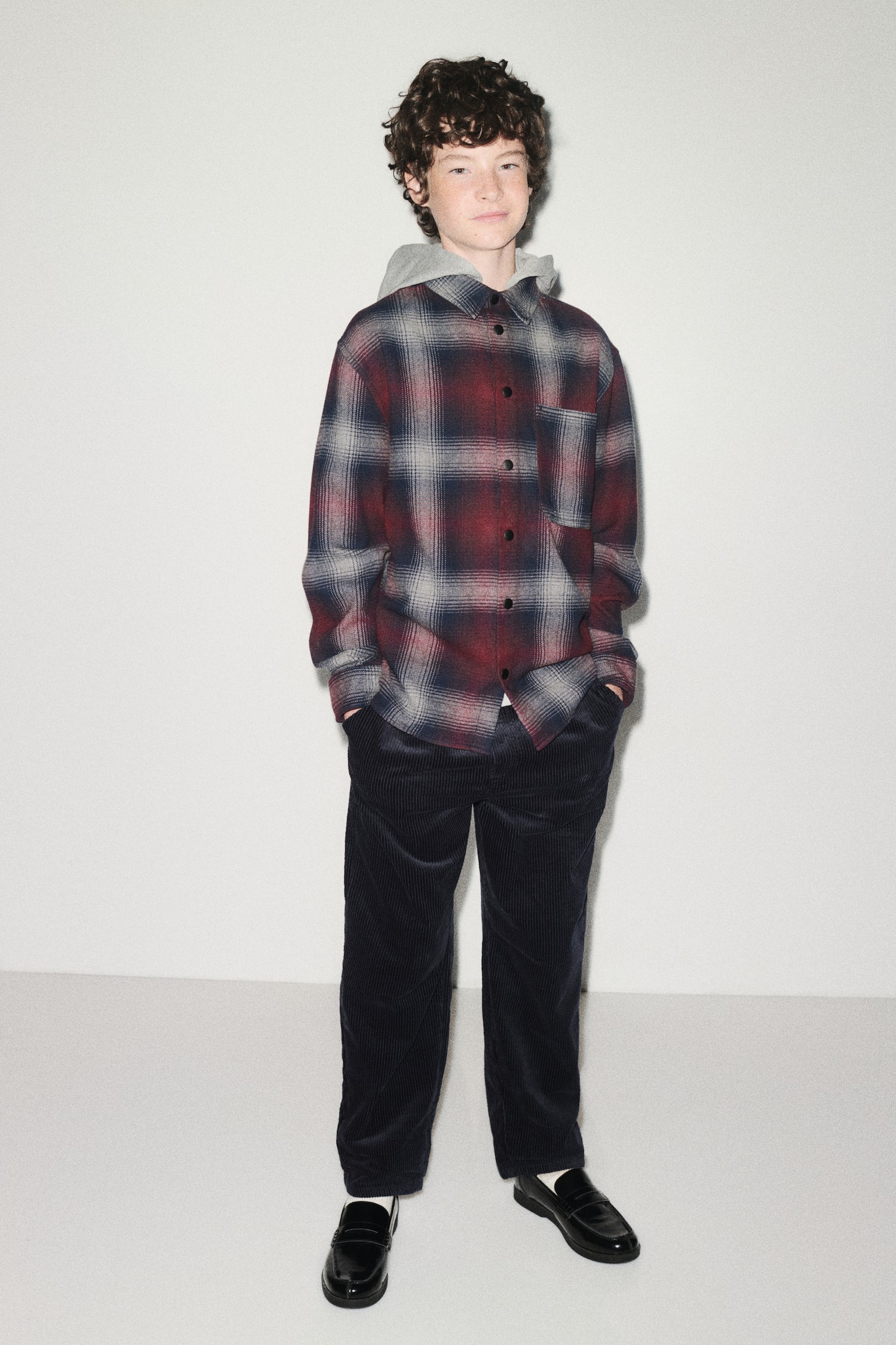 A loose-fit cotton flannel overshirt in a red, grey, and black plaid pattern is being worn facing the front, featuring a detachable light grey hood, a classic collar, and black press studs down the front. The overshirt displays a single open chest pocket and long sleeves with press-stud cuffs, layered over dark corduroy trousers. - 1