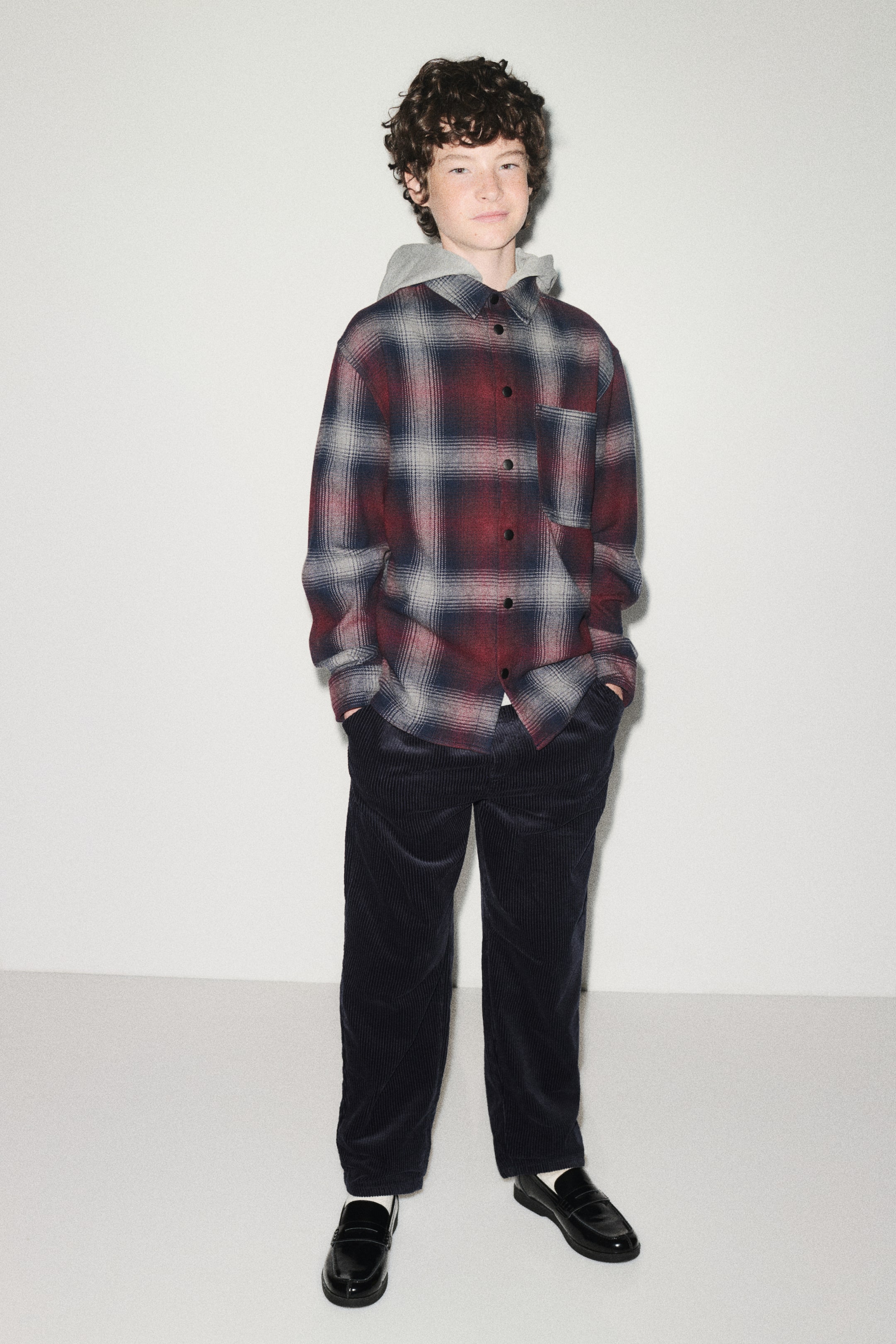 View larger image: A loose-fit cotton flannel overshirt in a red, grey, and black plaid pattern is being worn facing the front, featuring a detachable light grey hood, a classic collar, and black press studs down the front. The overshirt displays a single open chest pocket and long sleeves with press-stud cuffs, layered over dark corduroy trousers.