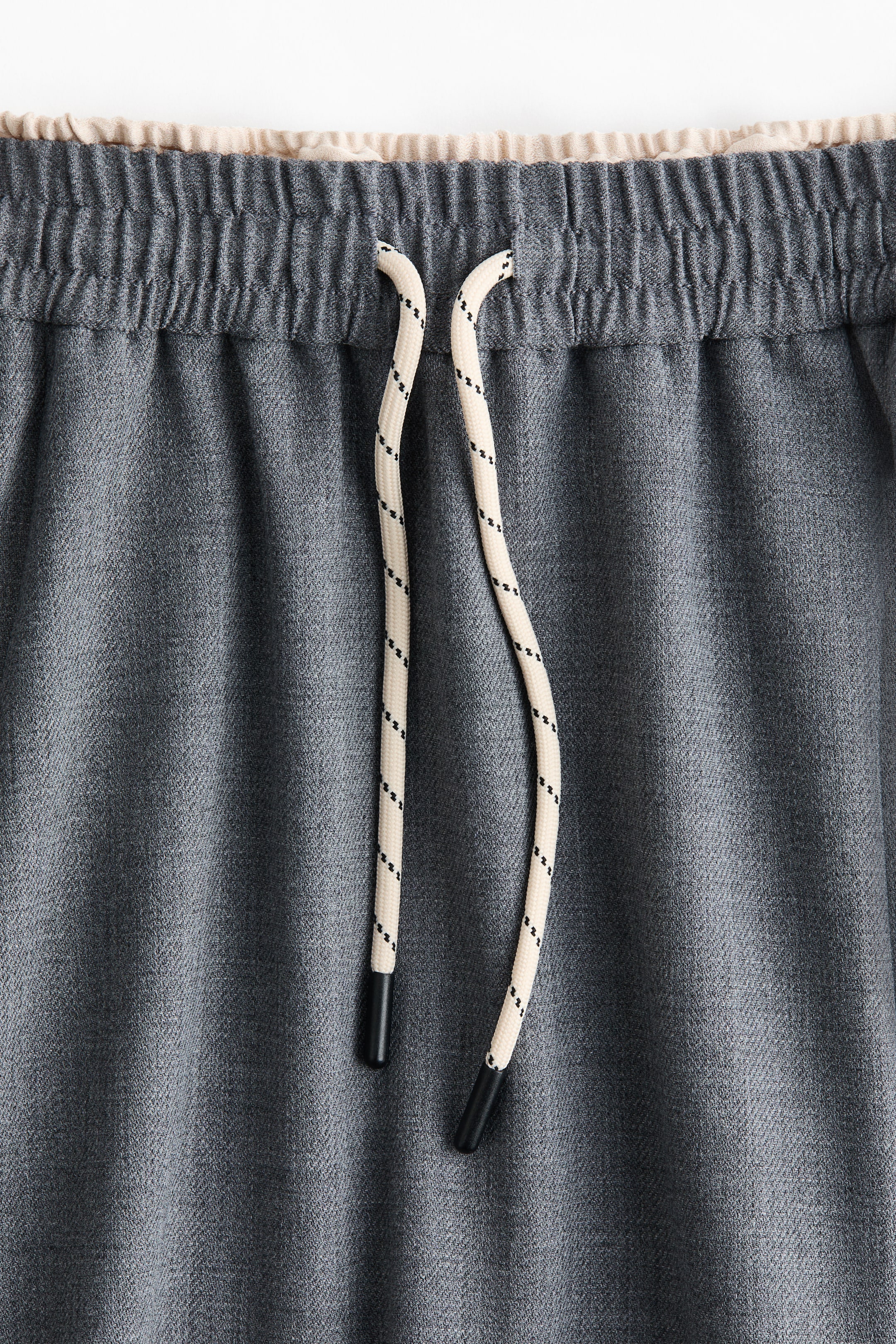View larger image: The front of dark gray drawstring pants is featuring an elasticated, gathered waistband with a contrasting light beige inner lining. Cream-colored drawstrings with black flecks and black tips are hanging from the front of the waistband.
