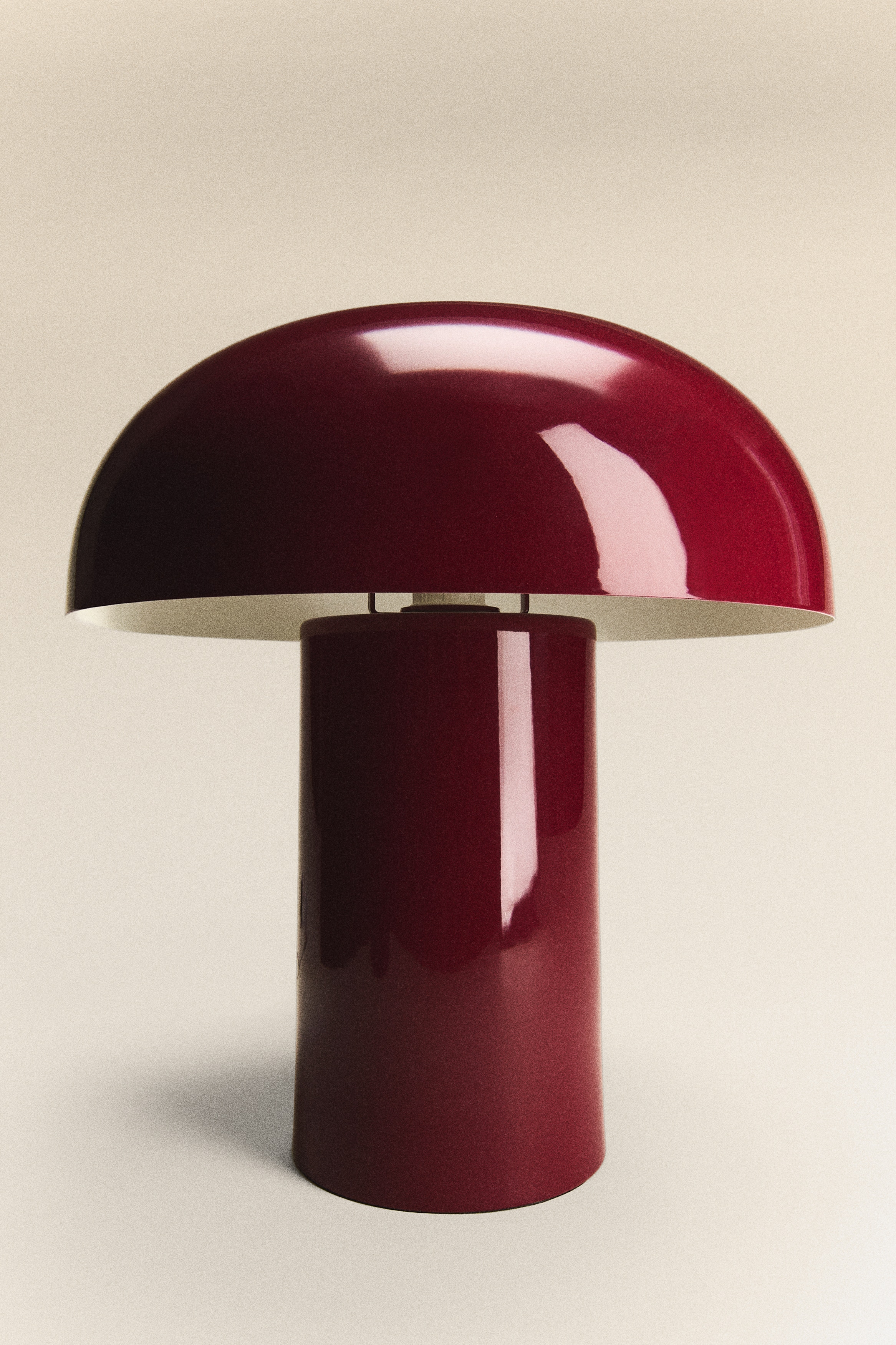 A large, deep red metal table lamp featuring a rounded, mushroom-inspired shape, presented from the front. A light grey, fabric-covered cord extends from the lamp's cylindrical base.