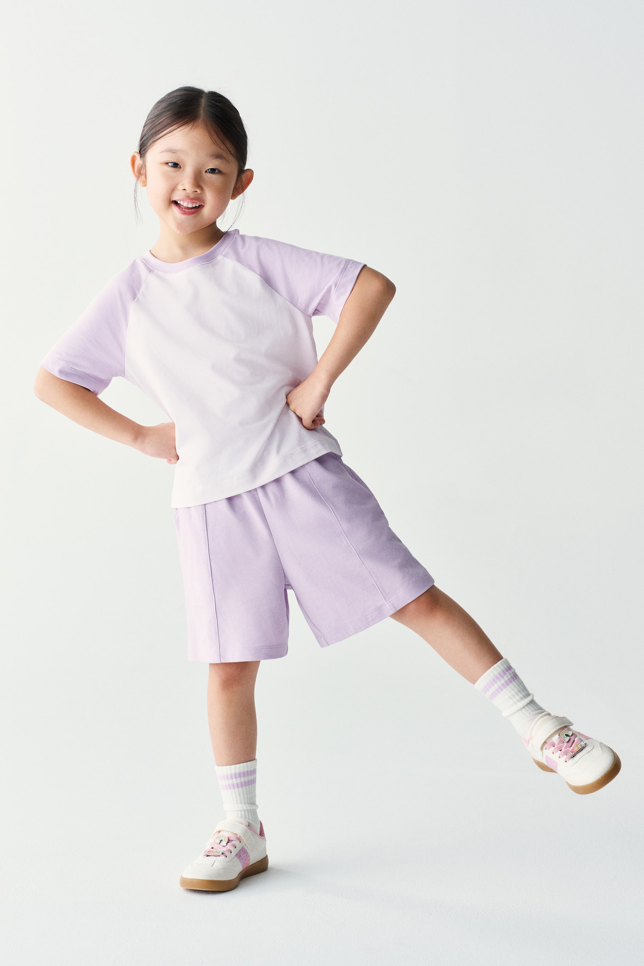 Xem ảnh lớn hơn: Một bộ đồ thể thao hai món gồm áo thun tay raglan màu tím nhạt và trắng cùng quần short tím nhạt đồng bộ đang được người mẫu mặc. Trang phục được phối cùng tất cổ ngắn sọc và giày sneaker trắng.