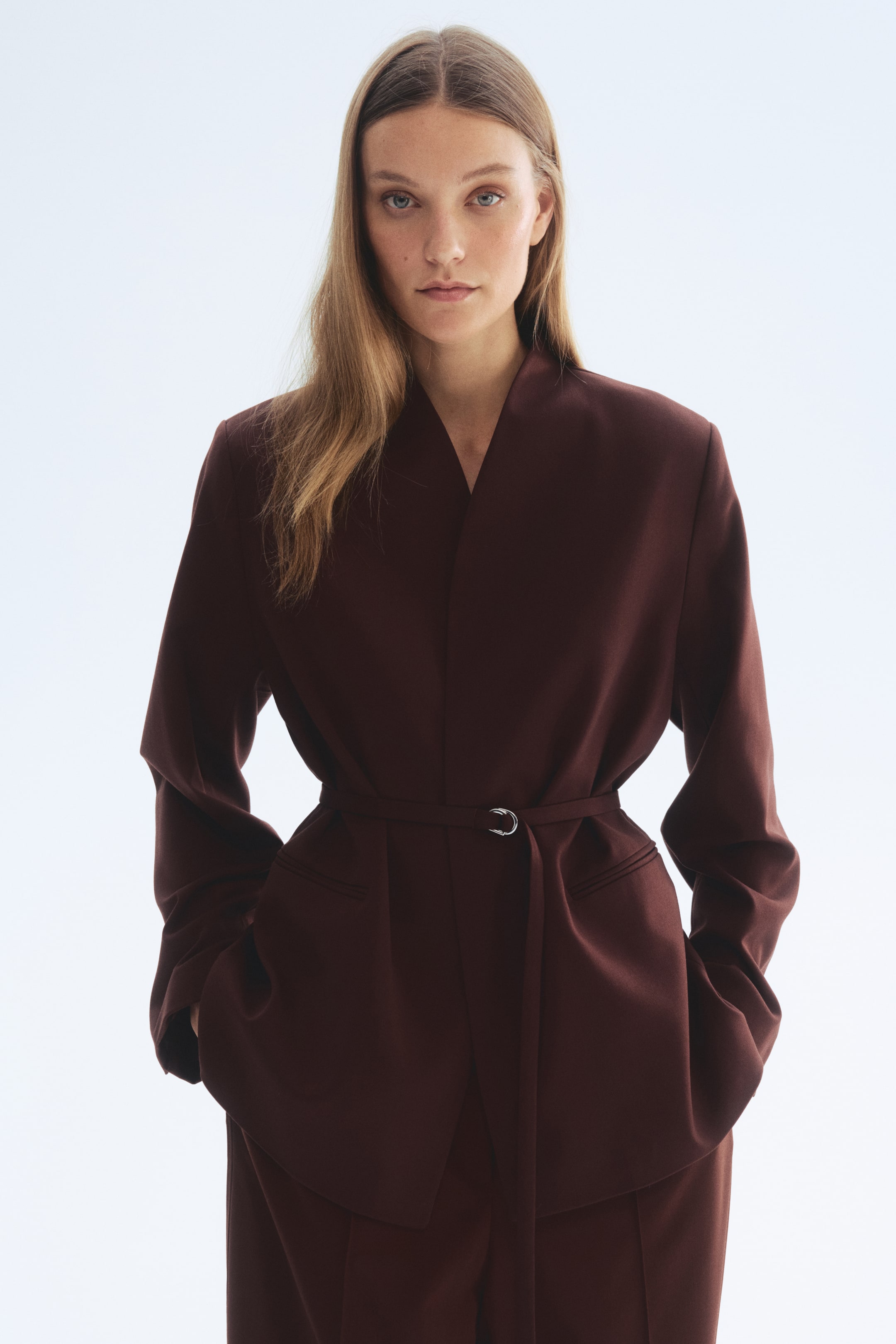 View larger image: FRONT view of an oversized blazer in dark reddish-brown woven fabric, featuring a stand collar, a wrapover front, and structured shoulder pads. A narrow belt with a silver ring buckle is cinching the waist, and it features long sleeves with slits at the cuffs and jetted front pockets.
