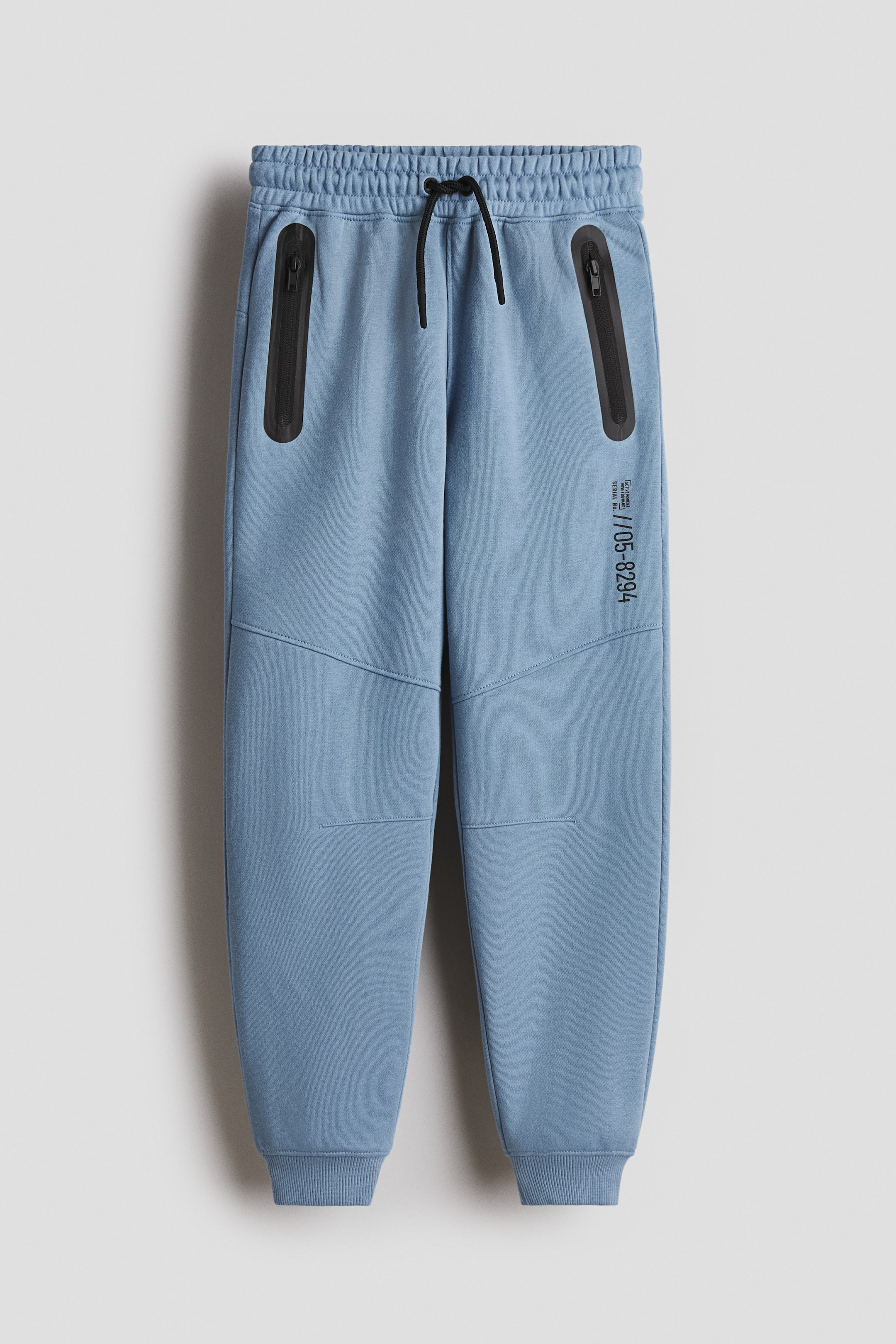 View larger image: A pair of blue sweatpants is shown FRONT, featuring an elasticated waistband with a black drawstring and two black vertical zip pockets on the upper front. Geometric seam details are visible on the legs, along with black vertical text on the right leg and ribbed cuffs at the ankles.