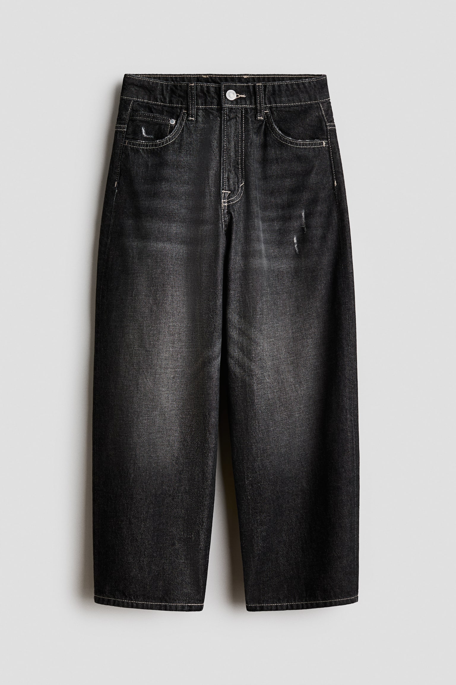 Baggy 5-pocket jeans van gewassen zwart katoenen denim hangen frontaal in beeld en tonen een wide-leg silhouet met lichte contraststiksels. De jeans heeft een vervaagde wassing over de bovenbenen, een klassieke sluiting met knoop en rits, en subtiele distressed details op het muntzakje en de linker bovenbeen.