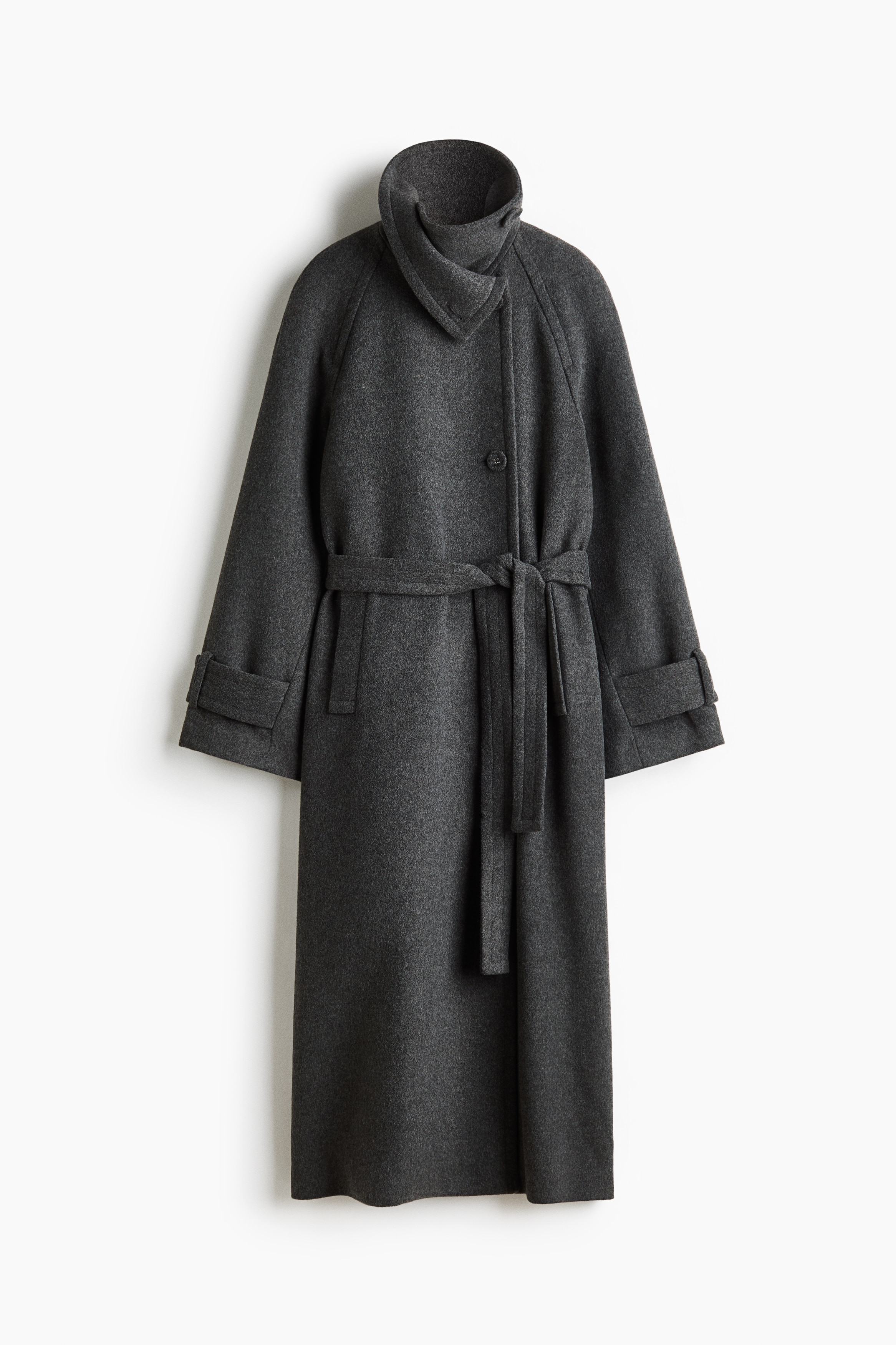 A dark grey felted maxi coat is shown from a side-back profile facing left, highlighting its long raglan sleeves with button-tab cuffs and a wide tie belt at the waist. The coat is layered over a light grey hoodie and wide-leg blue jeans, showcasing its long length and back vent.