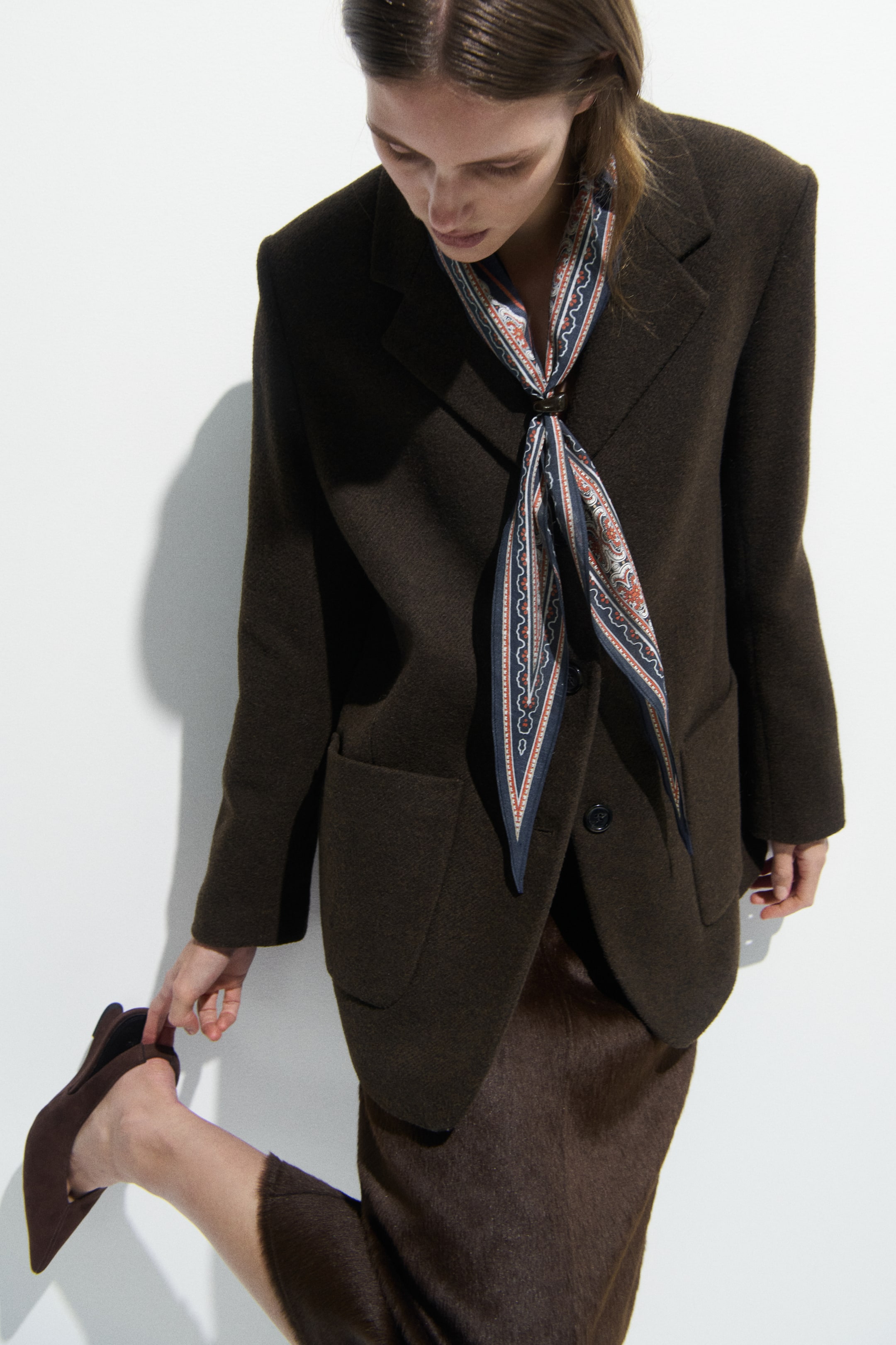 View larger image: A dark brown, single-breasted blazer is worn, featuring wide notched lapels and two large front patch pockets. A patterned scarf is styled over the blazer, secured with a ring at the neckline.