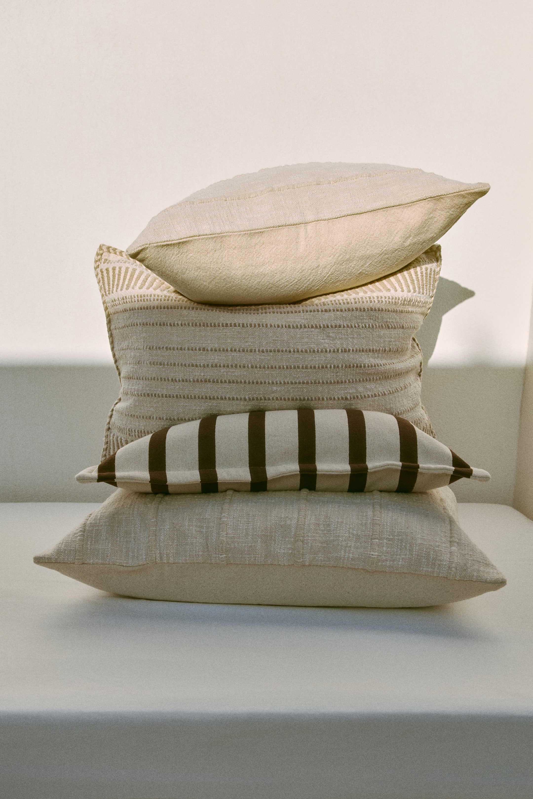 View larger image: A stack of four textured pillows showcases diverse shapes and patterns in neutral shades. The top light beige rectangular pillow rests on a larger rectangular pillow adorned with geometric designs and horizontal stripes. Beneath this, an elongated pillow with striking brown vertical stripes rests on the bottom light beige textured rectangular pillow.