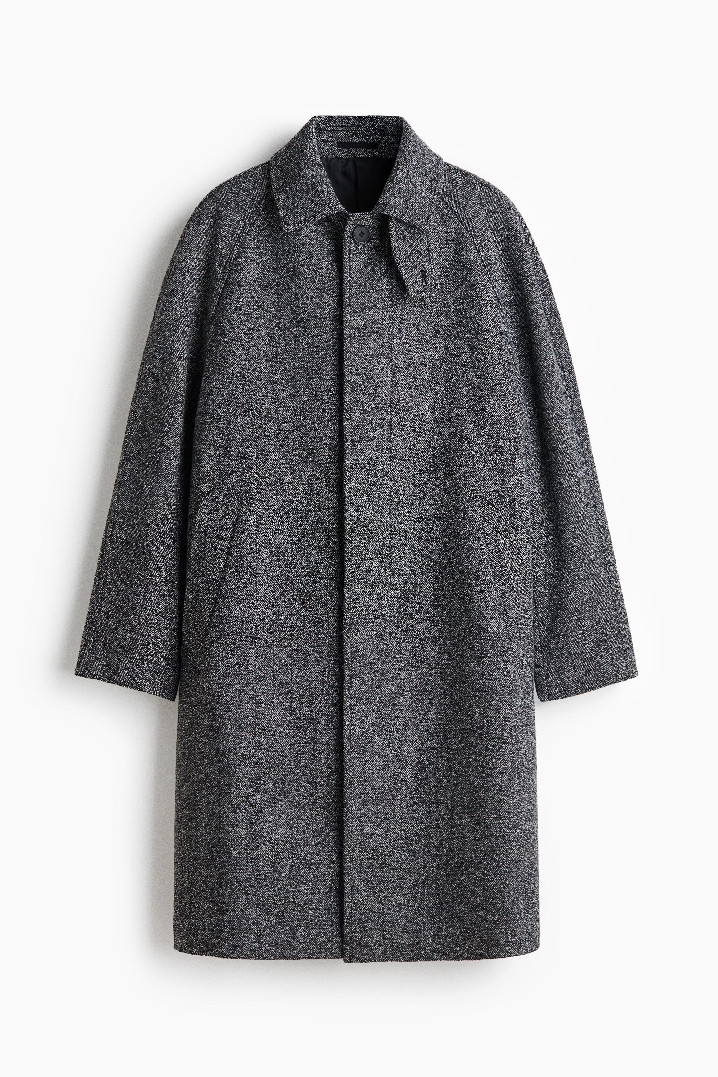 A knee-length car coat in a grey, herringbone-weave wool blend is worn by the model with one hand resting in the side pocket. The coat is styled open, showcasing its classic collar and raglan sleeves, layered over a dark blue zip-up jacket and charcoal grey trousers.