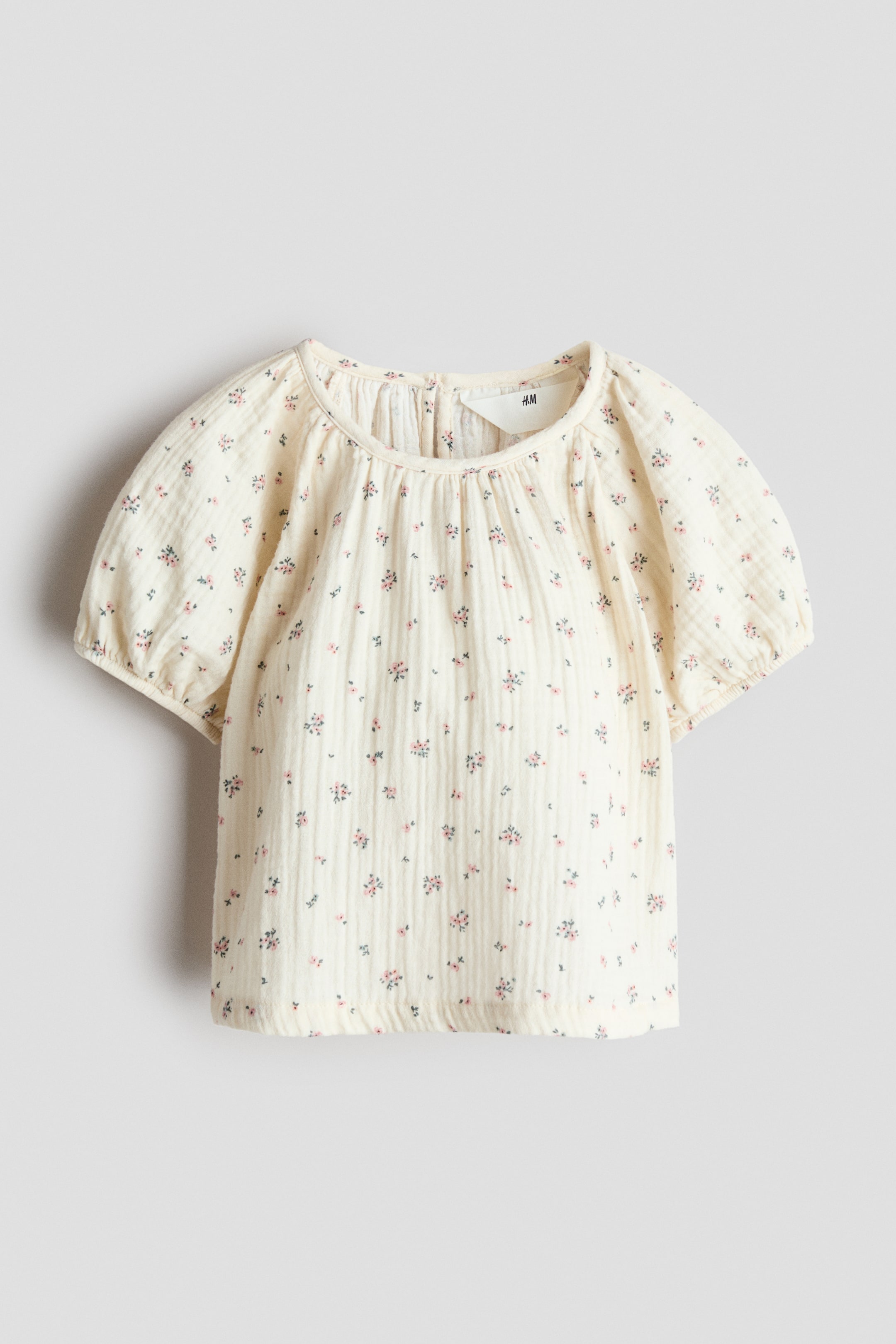 Ampliar la imagen: Se muestra de frente una blusa de muselina de algodón color crema con un pequeño estampado floral en rosa y verde. La blusa presenta un escote redondo fruncido y mangas cortas abullonadas con puños elásticos.