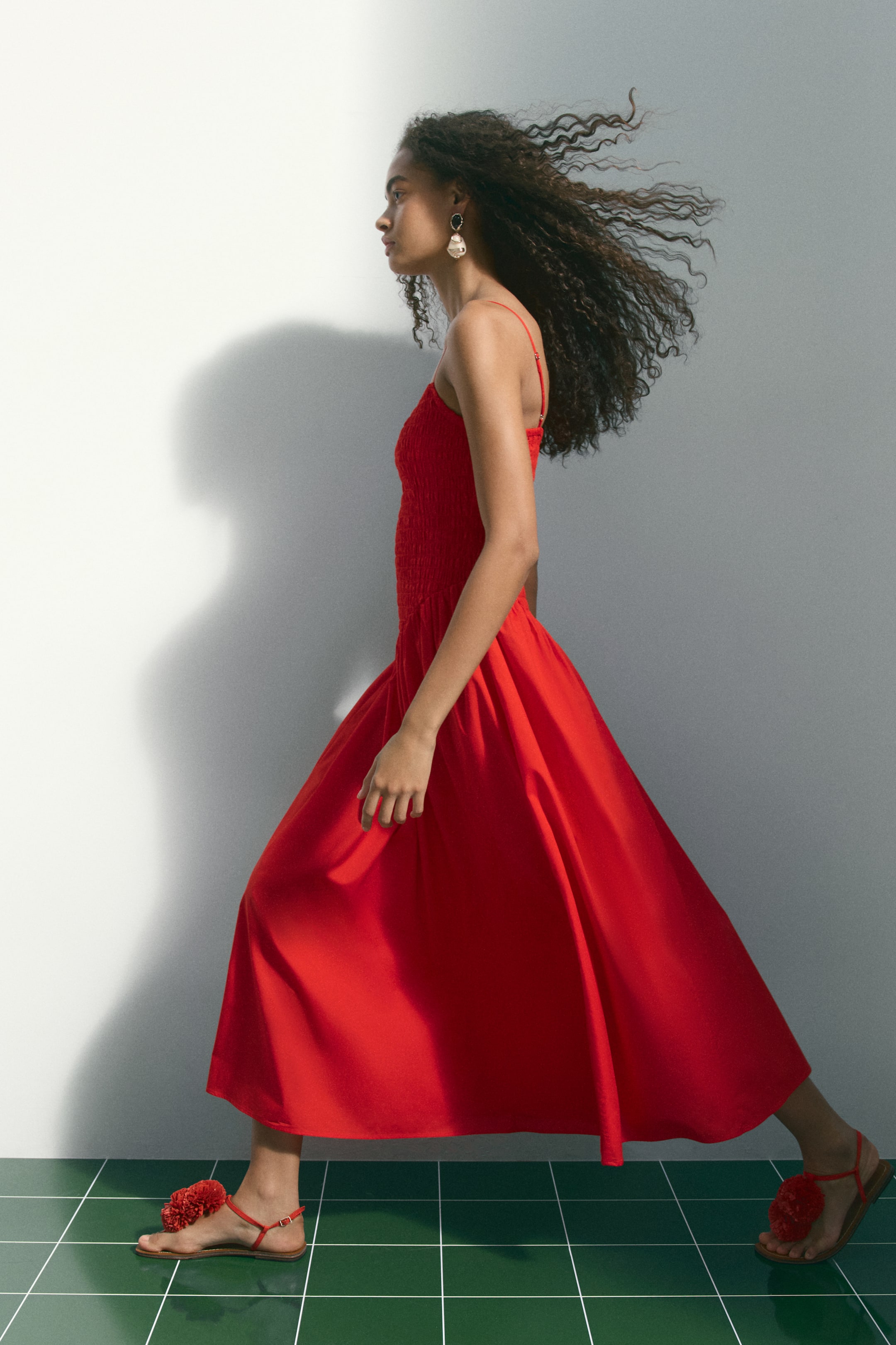 View larger image: A red midi dress menampilkan bodice berkerut dengan tali spaghetti dan skirt A-line yang mengalir, dilihat dari sisi. Ia dipadankan dengan sandal rata merah yang mempunyai hiasan pompom pada tali jari kaki.