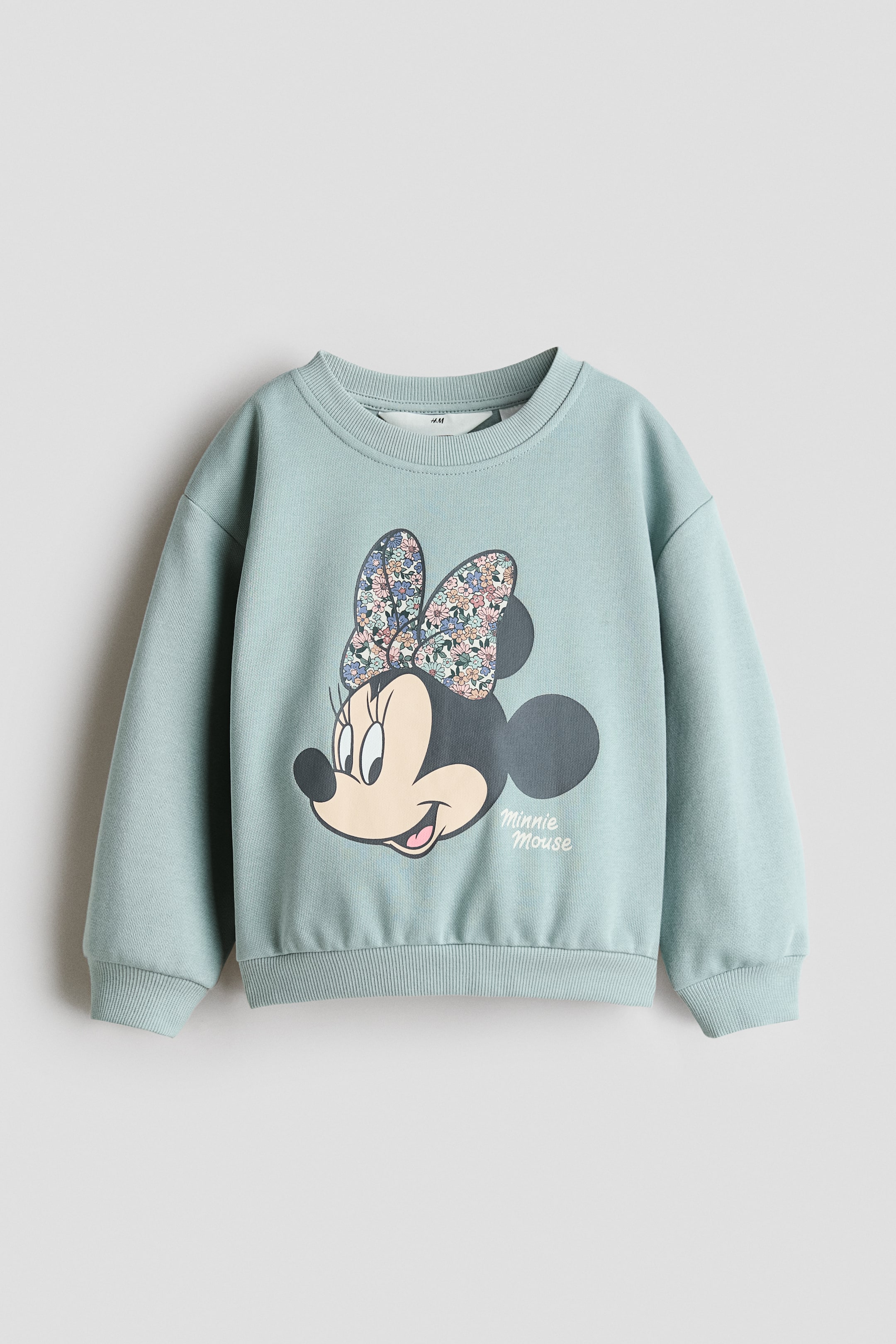 View larger image: A light blue sweatshirt is shown from the front, featuring a graphic print of Minnie Mouse's head with a floral bow and the text 'Minnie Mouse'. It is designed with a ribbed round neck, dropped shoulders, long sleeves with ribbed cuffs, and a ribbed hem.