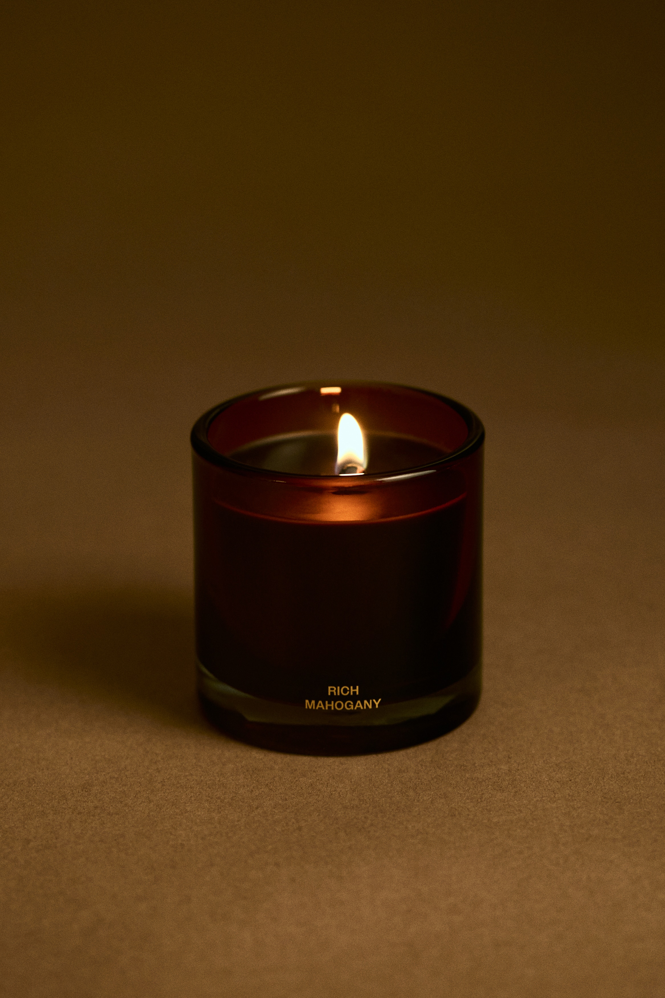 A scented candle in a cylindrical, amber-tinted glass jar is positioned upright and front-facing, containing dark wax with a single central wick. The jar features a thick, clear glass base and displays the gold-lettered text âRICH MAHOGANYâ near the lower front.