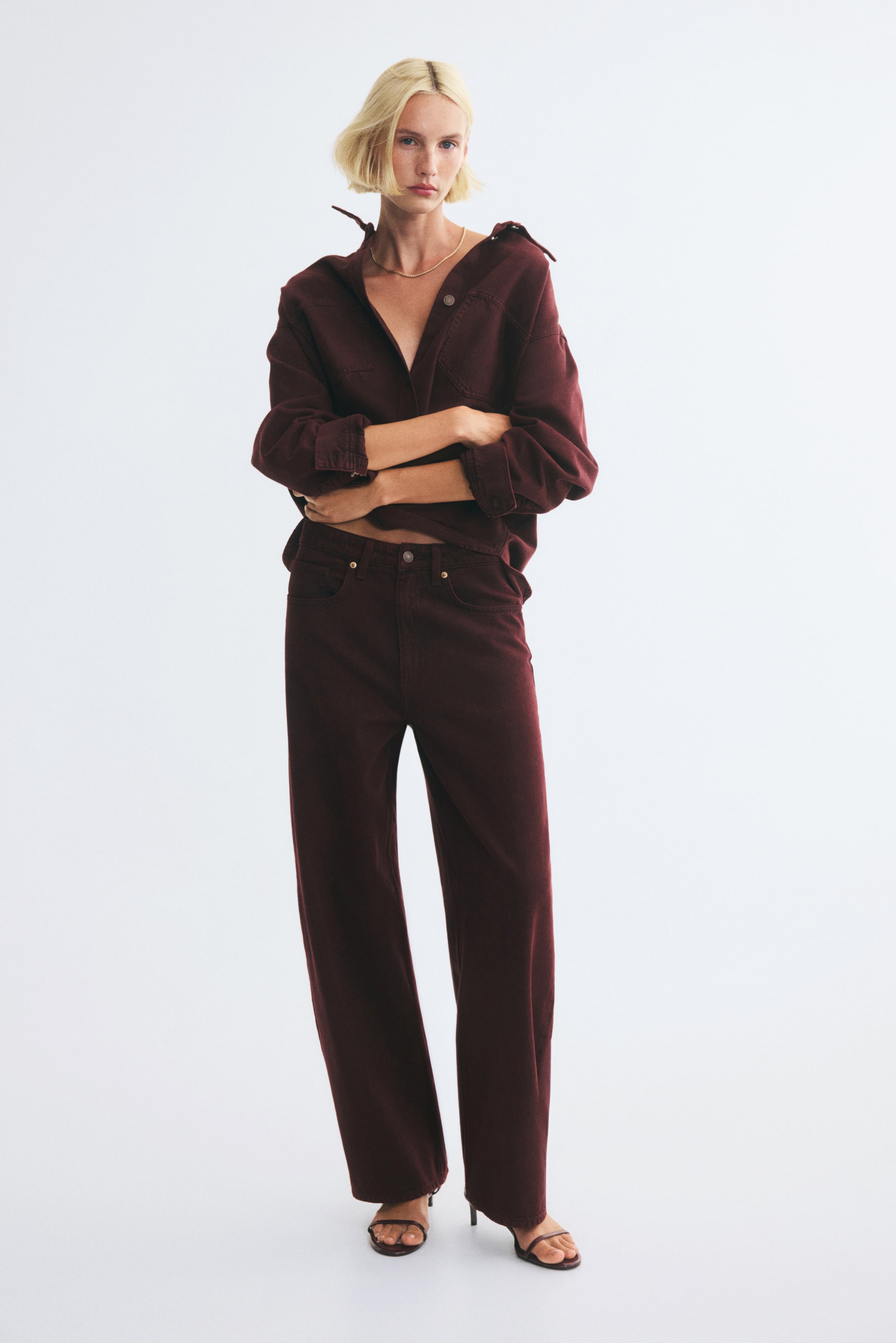 Grotere afbeelding bekijken: Een model draagt een high-waisted five-pocket jeans van donker bordeaux denim, met een gestructureerde regular fit die licht naar binnen loopt en smaller toeloopt naar de broekspijpen. De jeans wordt gecombineerd met een bijpassend bordeauxkleurig shirt met lange mouwen en donkere sandalen met bandjes en hoge hak.