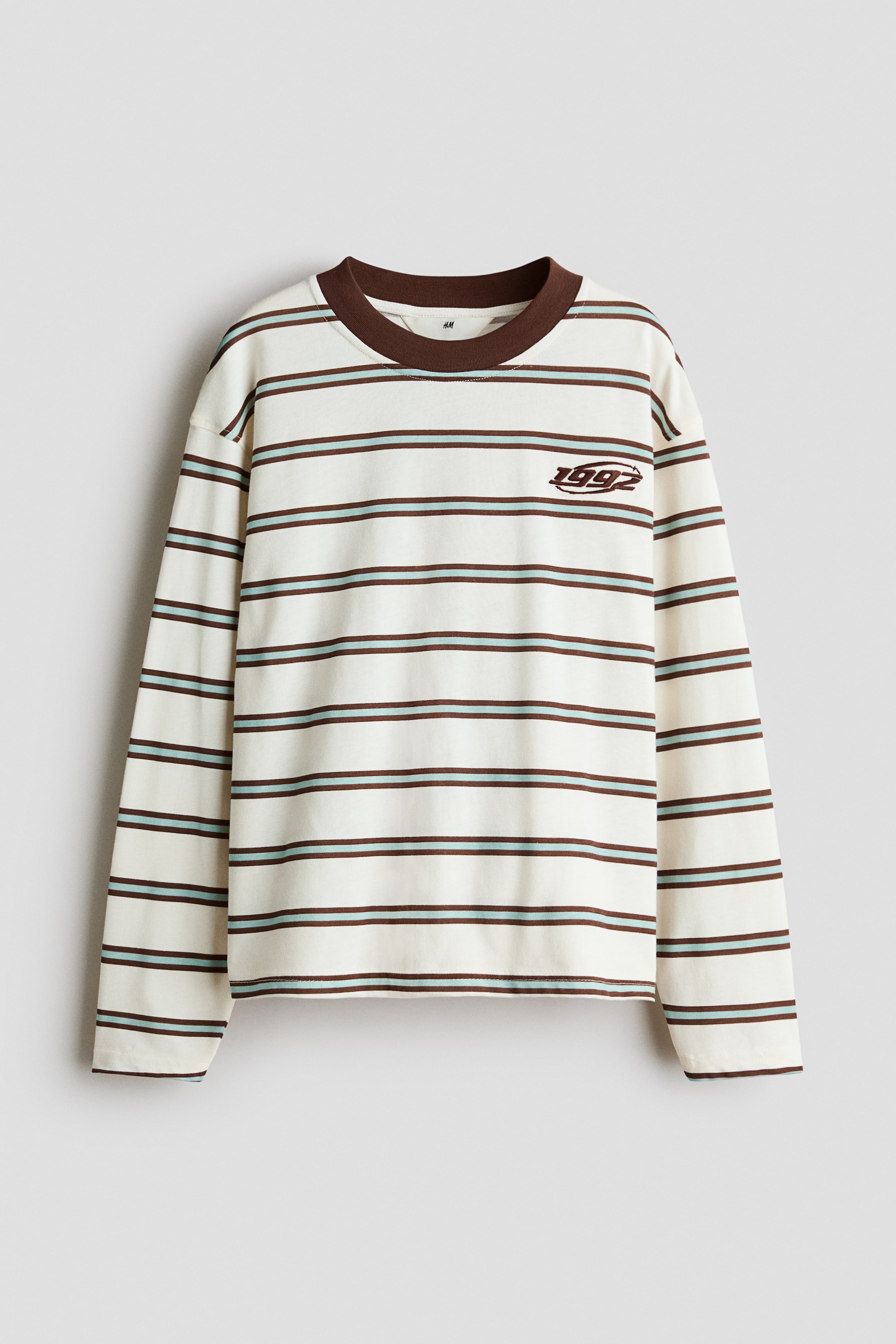 Visa större bild: En långärmad t-shirt visas framifrån, med en benvit bas och horisontella bruna och ljusblå ränder, en brun ribbad rund halsringning, och ett broderat "1992" på vänster bröst.