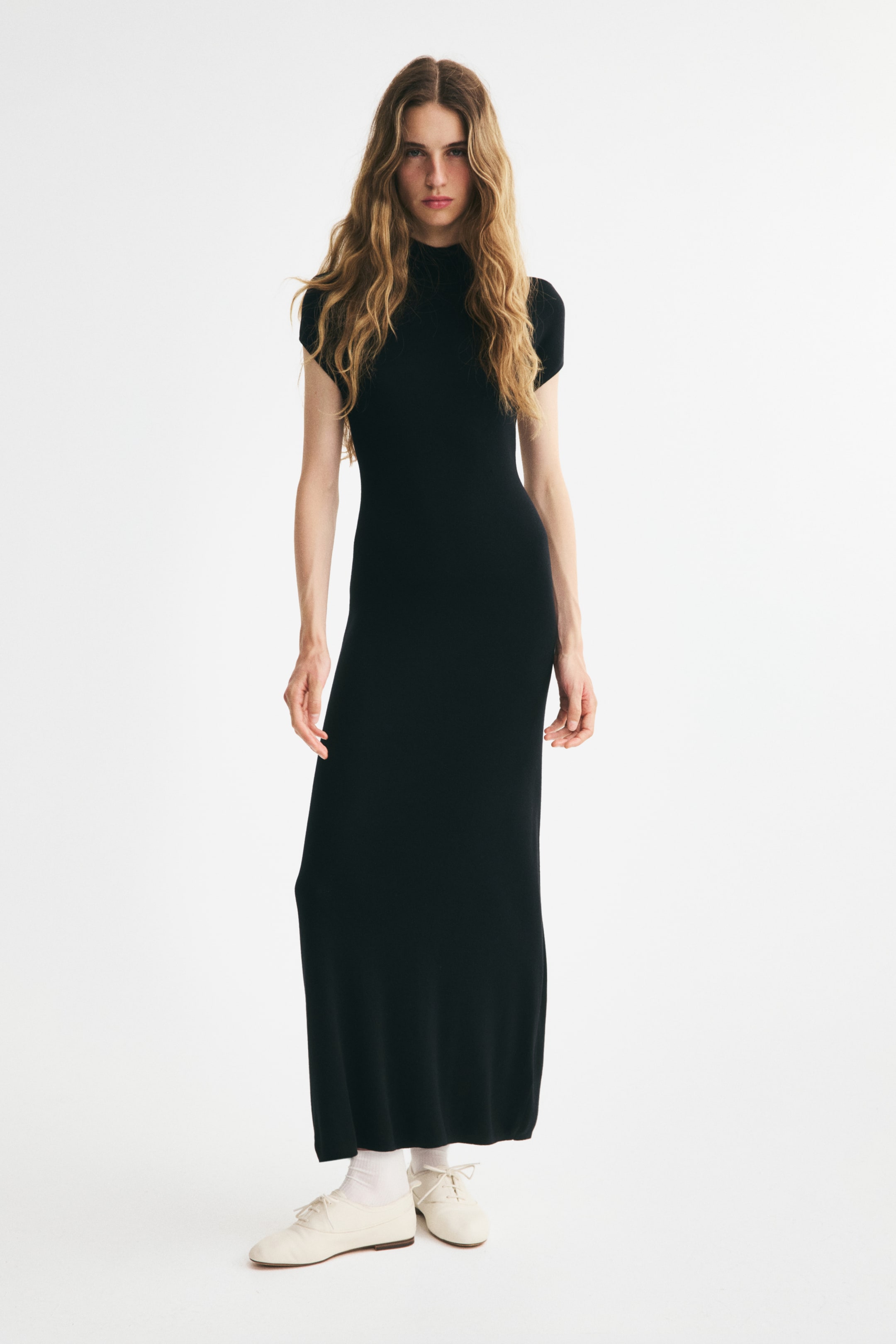 View larger image: A black knitted maxi dress is shown from the front, featuring a mock neck, short cap sleeves, and a figure-hugging silhouette. The dress falls straight to an ankle-length hem and is styled with white socks and cream-coloured lace-up shoes.