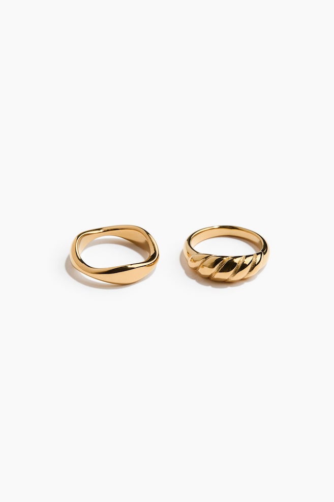 Two gold-toned rings rest side by side, facing forwards to showcase their polished finishes. The ring on the left features a smooth, organic wavy band, while the ring on the right highlights a textured, ribbed croissant-style design on its front. - 1