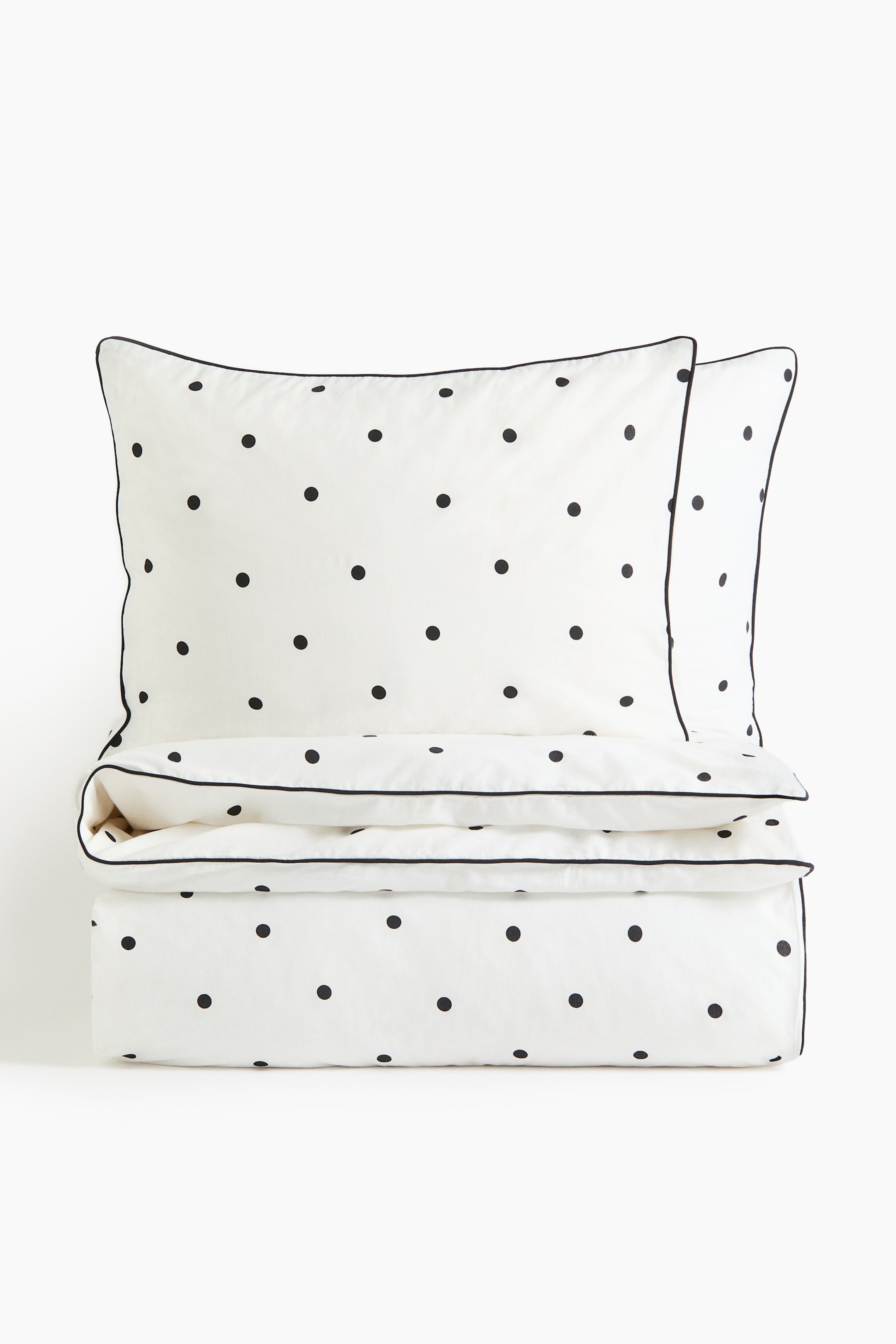 View larger image: A bedding set featuring a white fabric with an all-over black polka dot pattern and black piping along the edges. It showcases two pillowcases stacked at the top, with a folded duvet cover or sheet positioned beneath them.