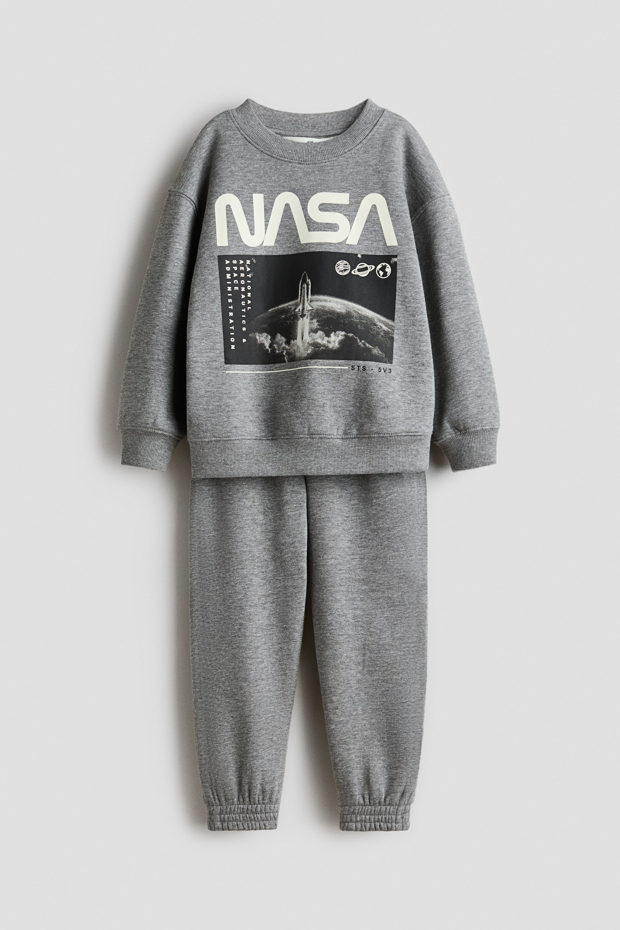 View larger image: A grey set, comprising a top and joggers in sweatshirt fabric, is being presented front-facing. The top is featuring a large graphic print on the front with 'NASA' lettering, a rocket launching from Earth, and small planetary icons, and is designed with a round, rib-trimmed neckline, dropped shoulders, and ribbed cuffs and hem. The joggers are designed with an elasticated waist and covered elastication at the hems.