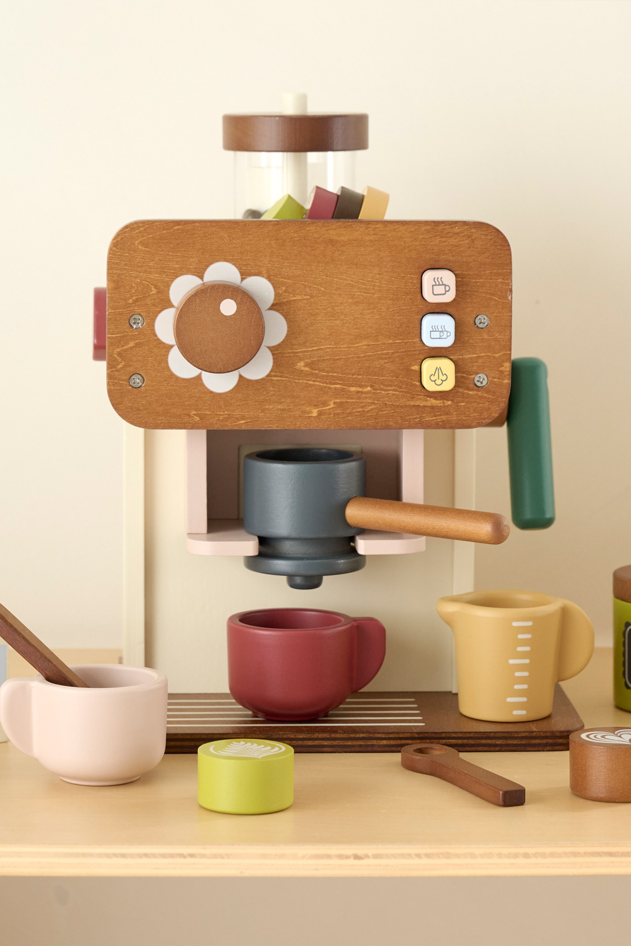 Agrandir l'image: Un ensemble de machine à café en bois est présenté de face, avec un corps marron, un cadran blanc en forme de fleur et trois boutons à icônes. Un porte-filtre gris est inséré dans la machine, tandis qu’une tasse rouge, une tasse rose avec un bâtonnet mélangeur, un pichet doseur jaune et des disques en bois avec latte art sont disposés à l’avant.
