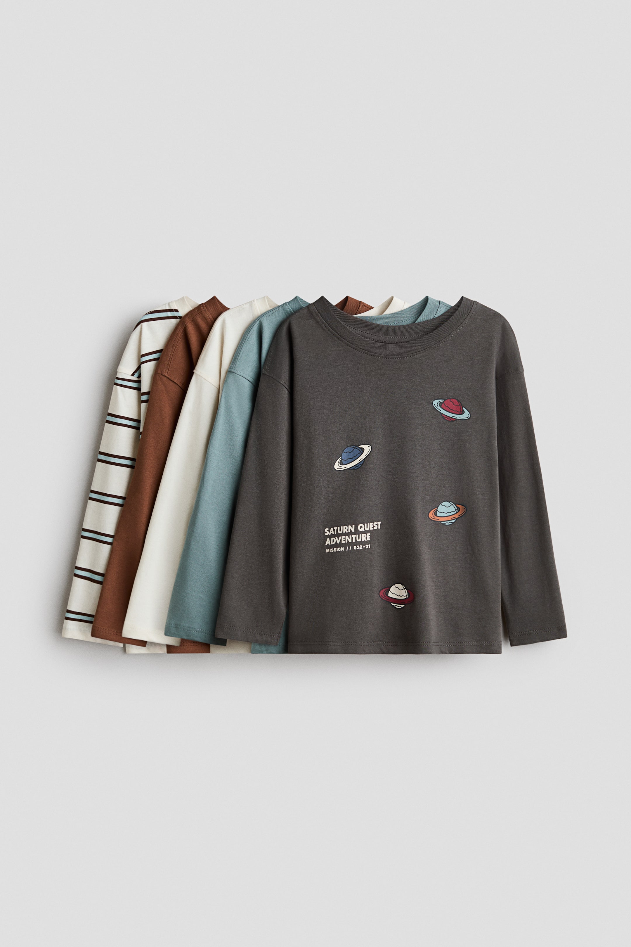 View larger image: Five long-sleeved t-shirts are layered, receding towards the left. The frontmost dark grey t-shirt showcases multiple vibrant planet illustrations and the text 'SATURN QUEST ADVENTURE' on its front. Behind it, you can see a solid teal, a solid cream, a solid brown, and a cream t-shirt with horizontal brown and light blue stripes.