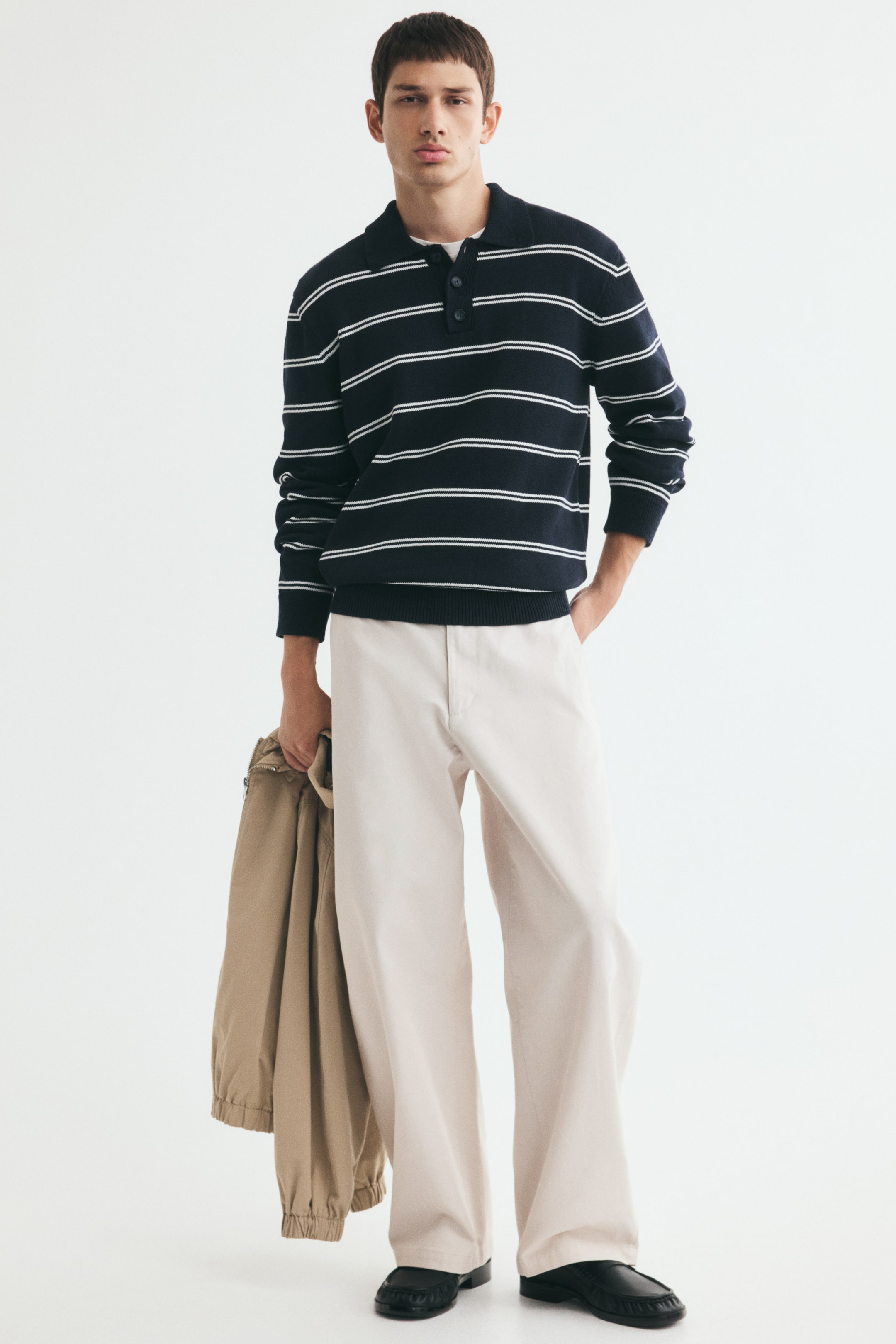 Agrandir l'image: Chino en coton de couleur crème, à coupe ample et jambes fuselées en forme de barrel, portés par le mannequin, qui se tient avec une main dans la poche de couture latérale. Le pantalon est associé à un polo en maille rayé bleu marine et blanc et à des mocassins noirs, tandis qu’une veste beige est tenue dans l’autre main du mannequin.