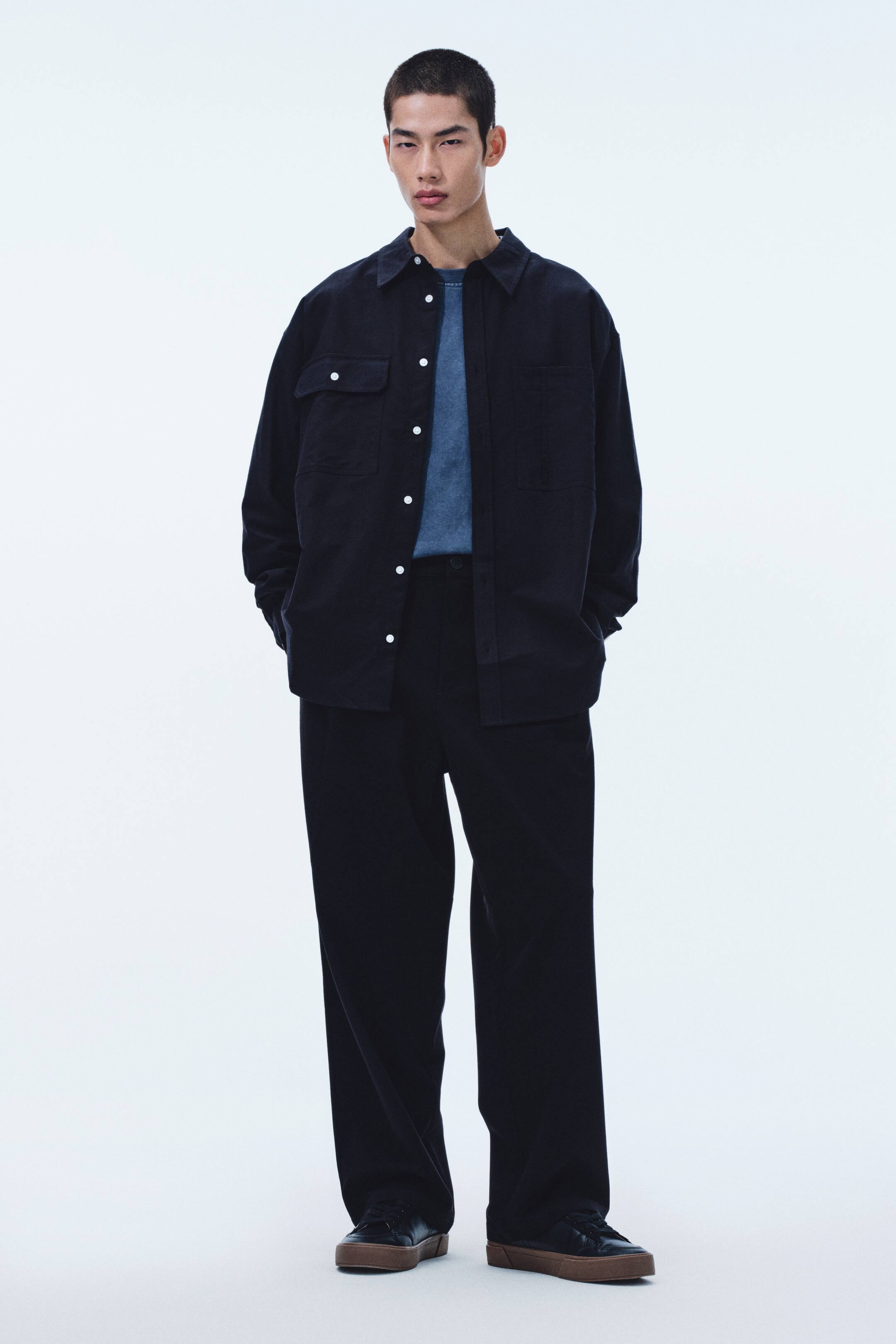 이미지 확대 보기: 다크 네이비 오버셔츠를 착용하고 있으며, 칼라와 흰색 버튼의 앞면 여밈, 가슴 플랩 포켓 2개 및 하단 패치 포켓 2개가 특징입니다. 페이드 블루 크루넥 티셔츠 위에 레이어드되었고, 같은 다크 네이비 와이드 팬츠와 갈색 밑창의 검정 스니커즈와 매치했습니다.