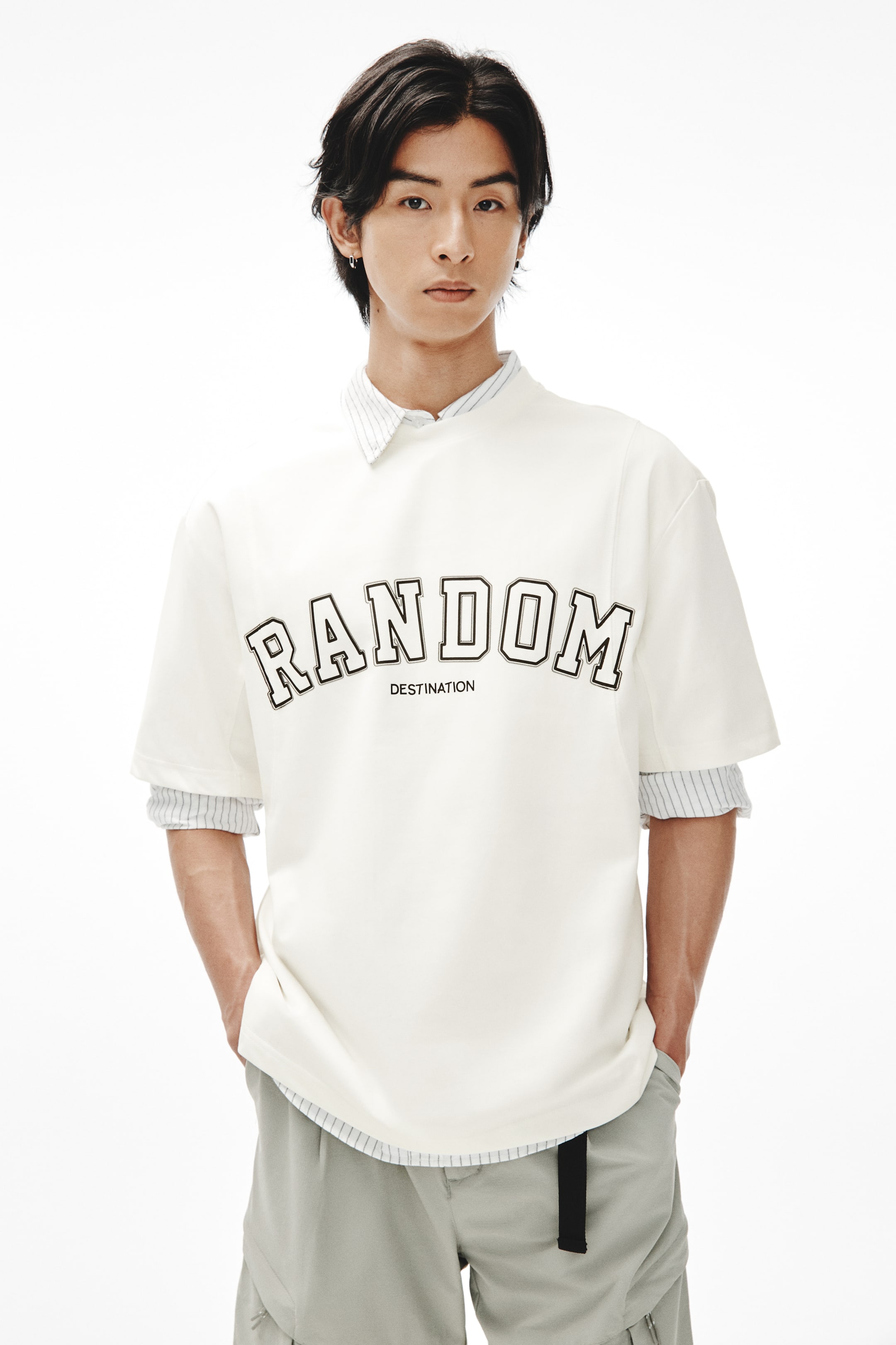 이미지 확대 보기: 모델이 입고 있는 크림색 반소매 셔츠는 "DESTINATION" 텍스트와 함께 "RANDOM" 그래픽이 윤곽으로 표시되어 있습니다. 이 셔츠는 밝은 색상의 줄무늬 칼라 셔츠 위에 레이어드되어 있으며, 칼라와 커프스가 보입니다.