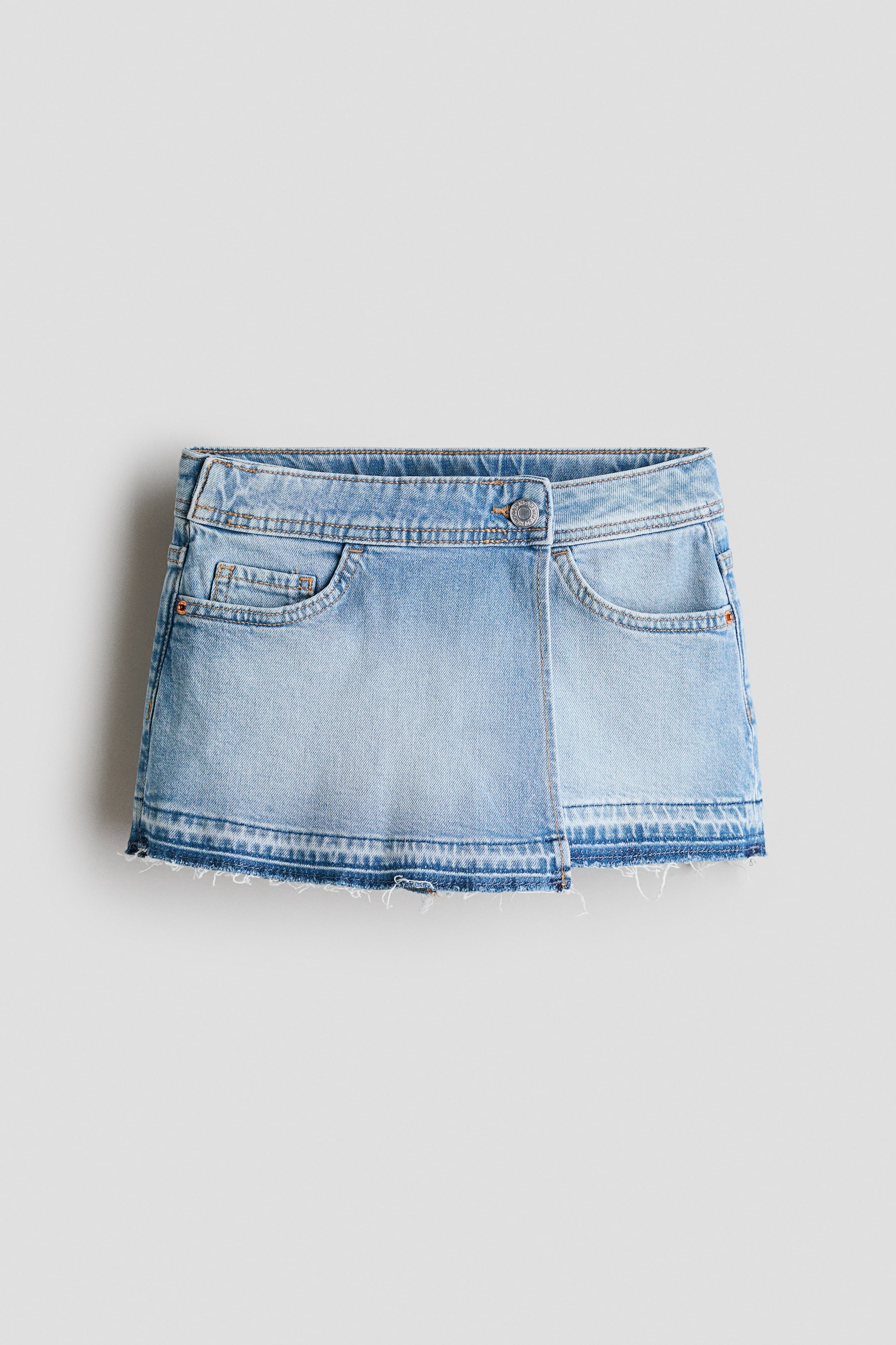 View larger image: A light blue denim skort is shown from the front, featuring an elasticated waistband. It displays a wrapover front with a silver-toned button fastening, two rounded front pockets, and a frayed hem.