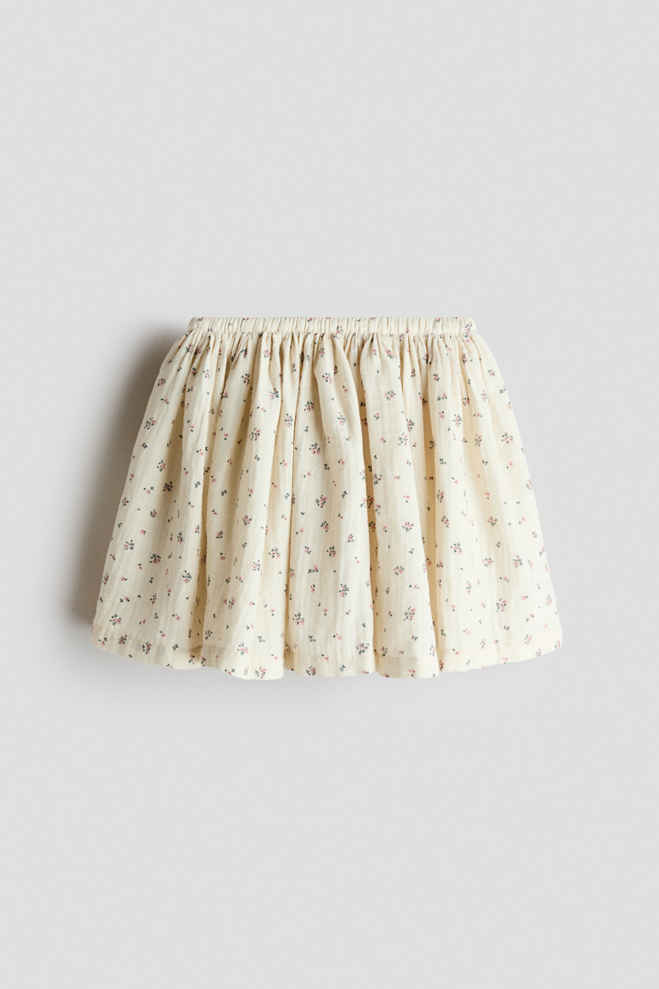 View larger image: The skirt is shown from the front, featuring a textured, cream-coloured fabric with a delicate, scattered floral print in pink and dark blue. Its elasticated, gathered waistband flows into a flared silhouette.