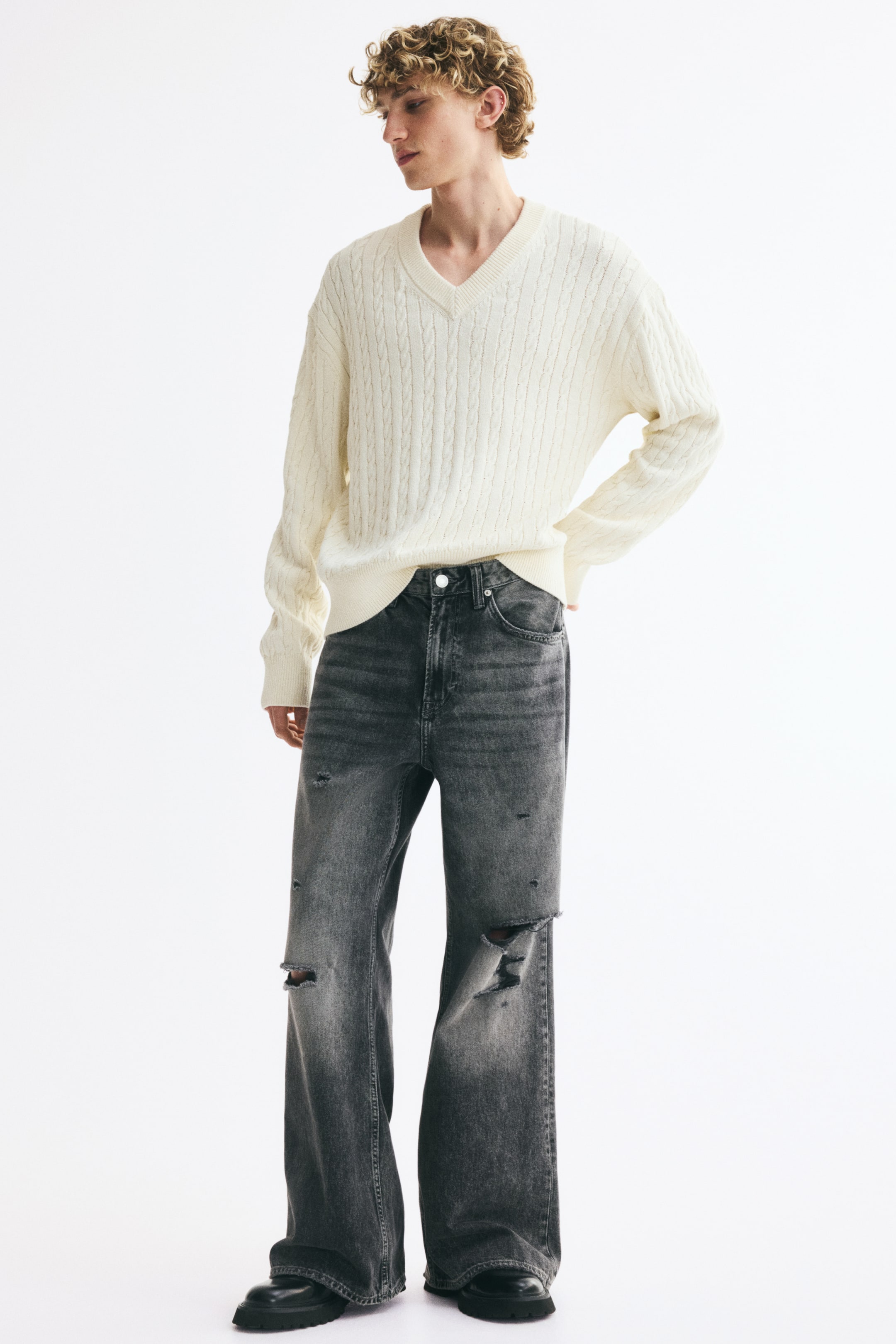 Agrandir l'image: Jean 5 poches gris foncé, confectionné en denim de coton rigide avec une coupe ample des hanches jusqu’à l’ourlet et une jambe légèrement évasée, porté par le mannequin qui glisse une main dans une poche avant. Le jean présente une taille standard avec fermeture zippée et bouton, ainsi que des détails usés sur les cuisses et les genoux.