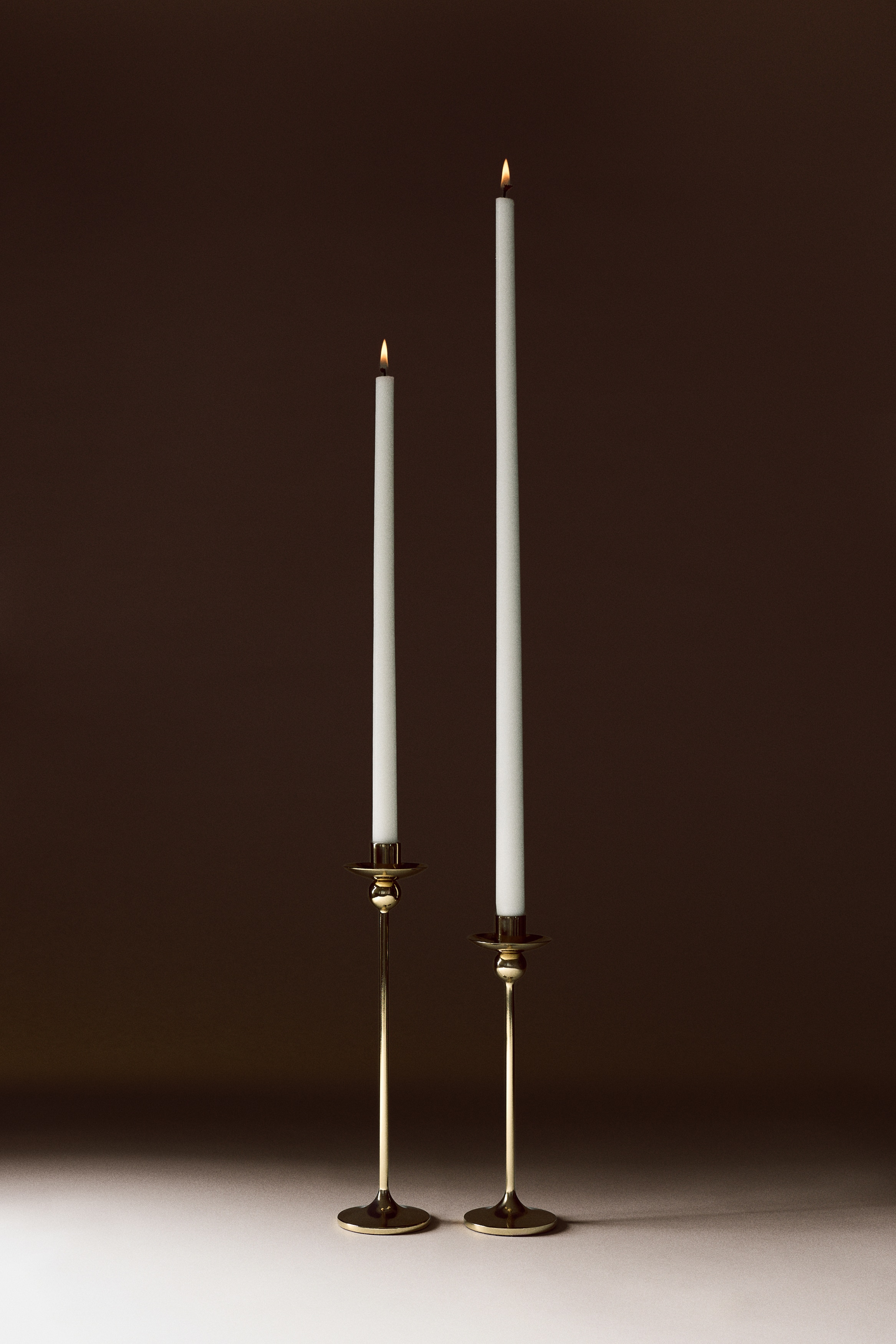 Un grand chandelier Ã  la finition dorÃ©e polie est prÃ©sentÃ© de face, avec une tige Ã©lÃ©gante et Ã©lancÃ©e sâÃ©levant depuis une base circulaire Ã©vasÃ©e. Le design comprend un dÃ©tail sphÃ©rique placÃ© juste en dessous dâune large coupelle Ã  cire peu profonde et un porte-bougie cylindrique au sommet.