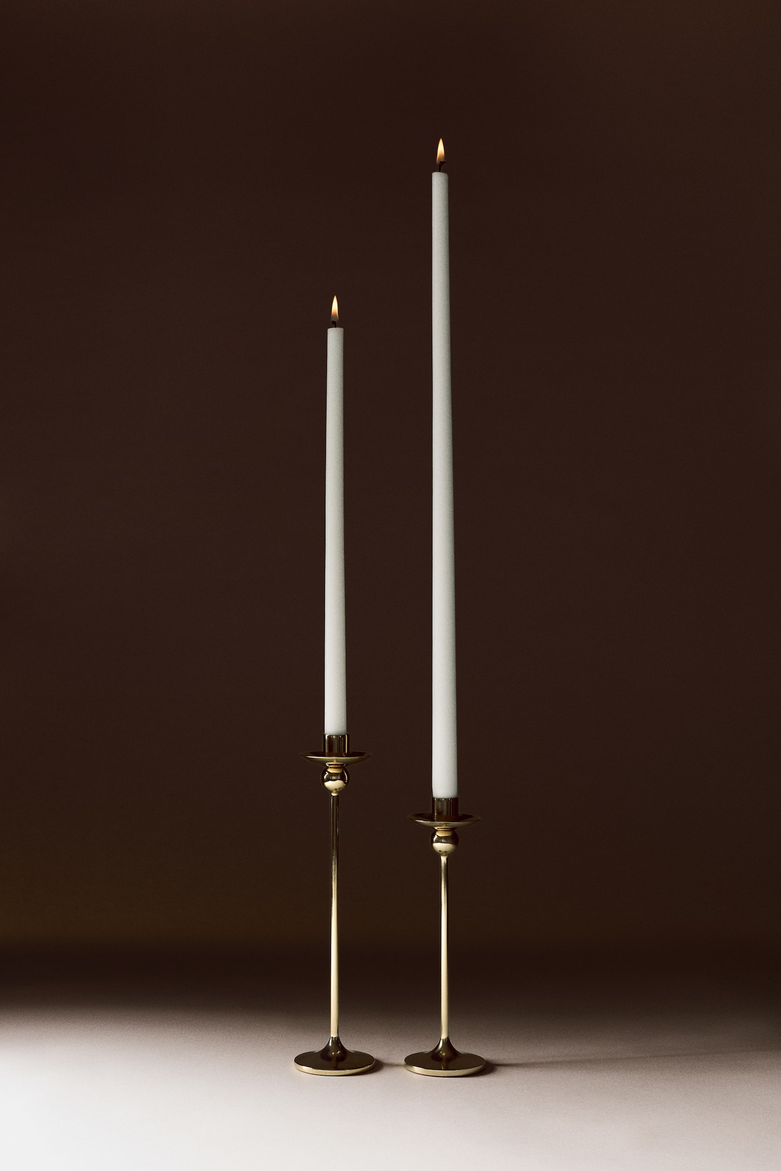 Tall candlesticks in polished gold-toned stainless steel are standing side-by-side, featuring a sleek, slender stem, a flat circular base, and a spherical detail just below the cup. Each candlestick is holding a lit, tall white taper candle. - 1