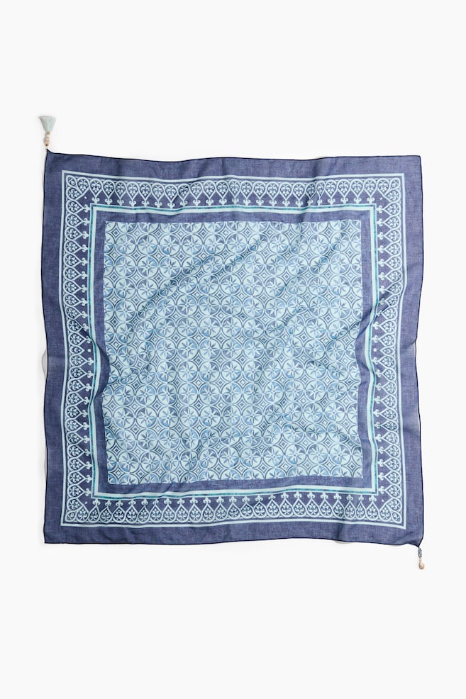A square scarf is lying flat, presenting its front side. It is featuring a central geometric pattern of circles and squares in shades of light blue, which is framed by a thin turquoise line, an intricate border of white heart-like motifs, and a wide outer border in dark blue. Small tassels are visible on two opposite corners. - 1